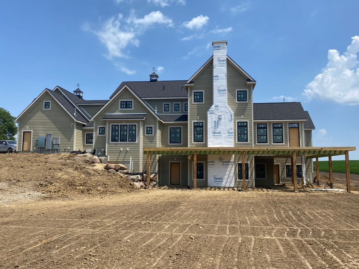 A large, light-colored house under construction with a wraparound wooden deck and an unfinished fireplace chimney.