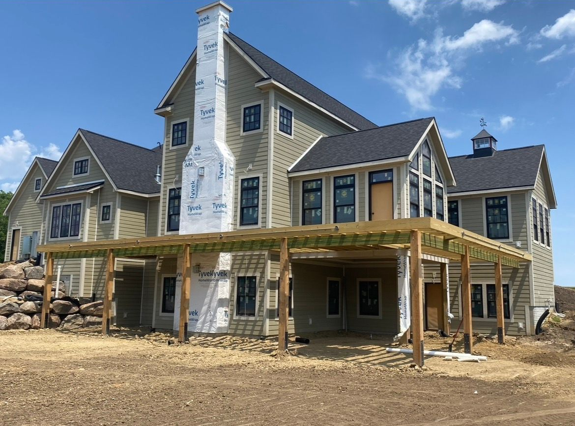 A large two-story house under construction with beige siding, a dark roof, and a wooden pergola over an unfinished patio.