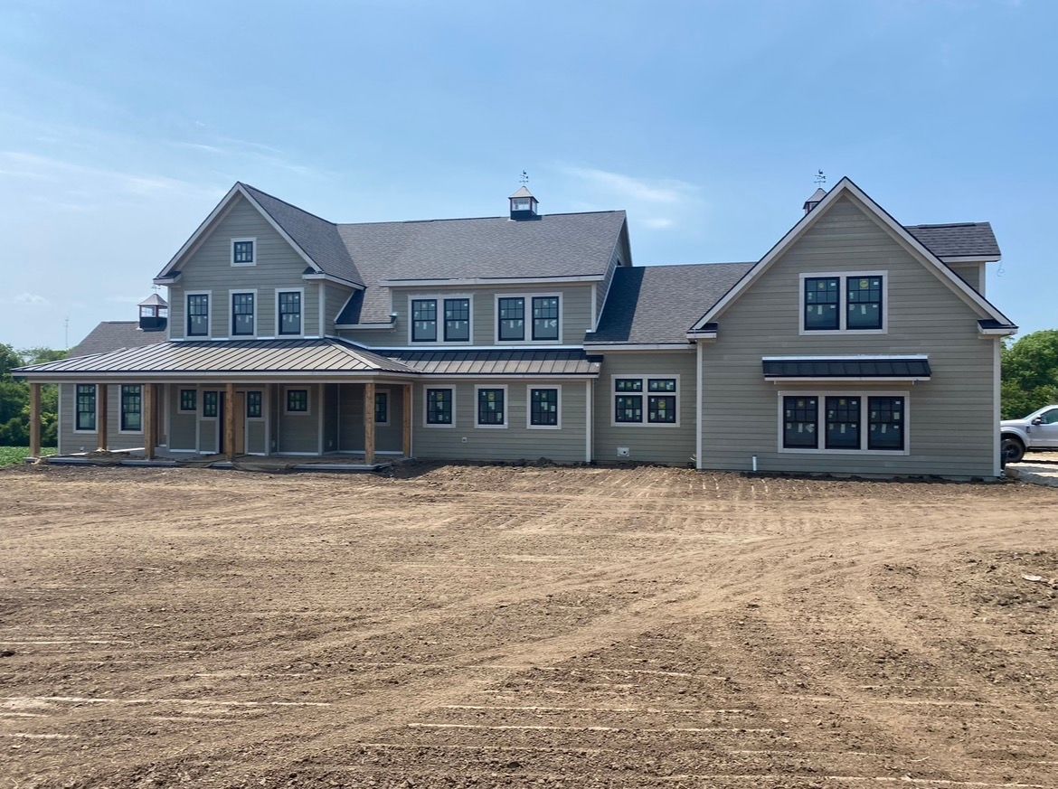 A large, light-gray two-story house under construction with a wraparound porch and dark roof, set on a bare dirt lot.