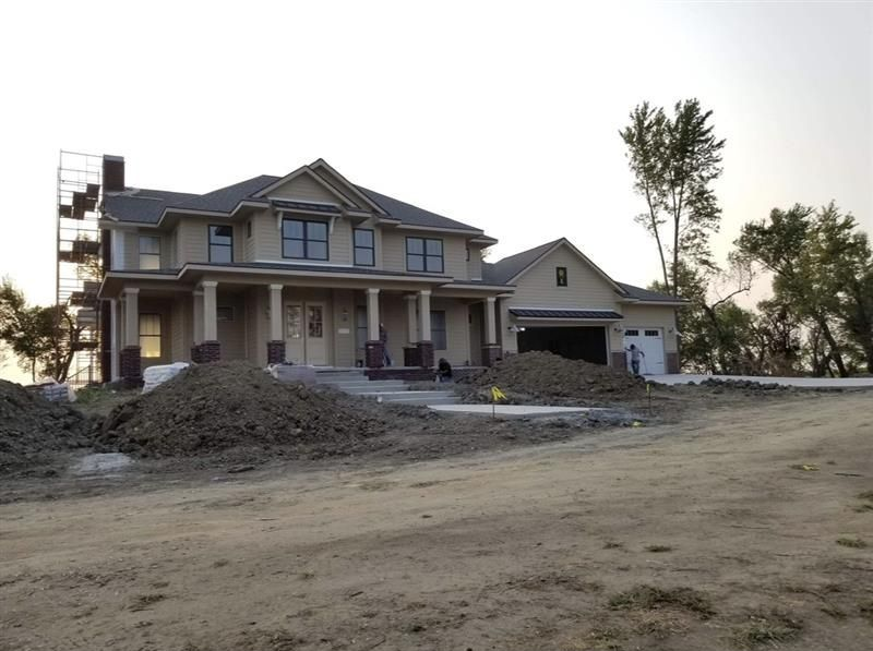 A two-story tan house under construction with a front porch, garage, and scaffolding, set in an unfinished dirt yard.