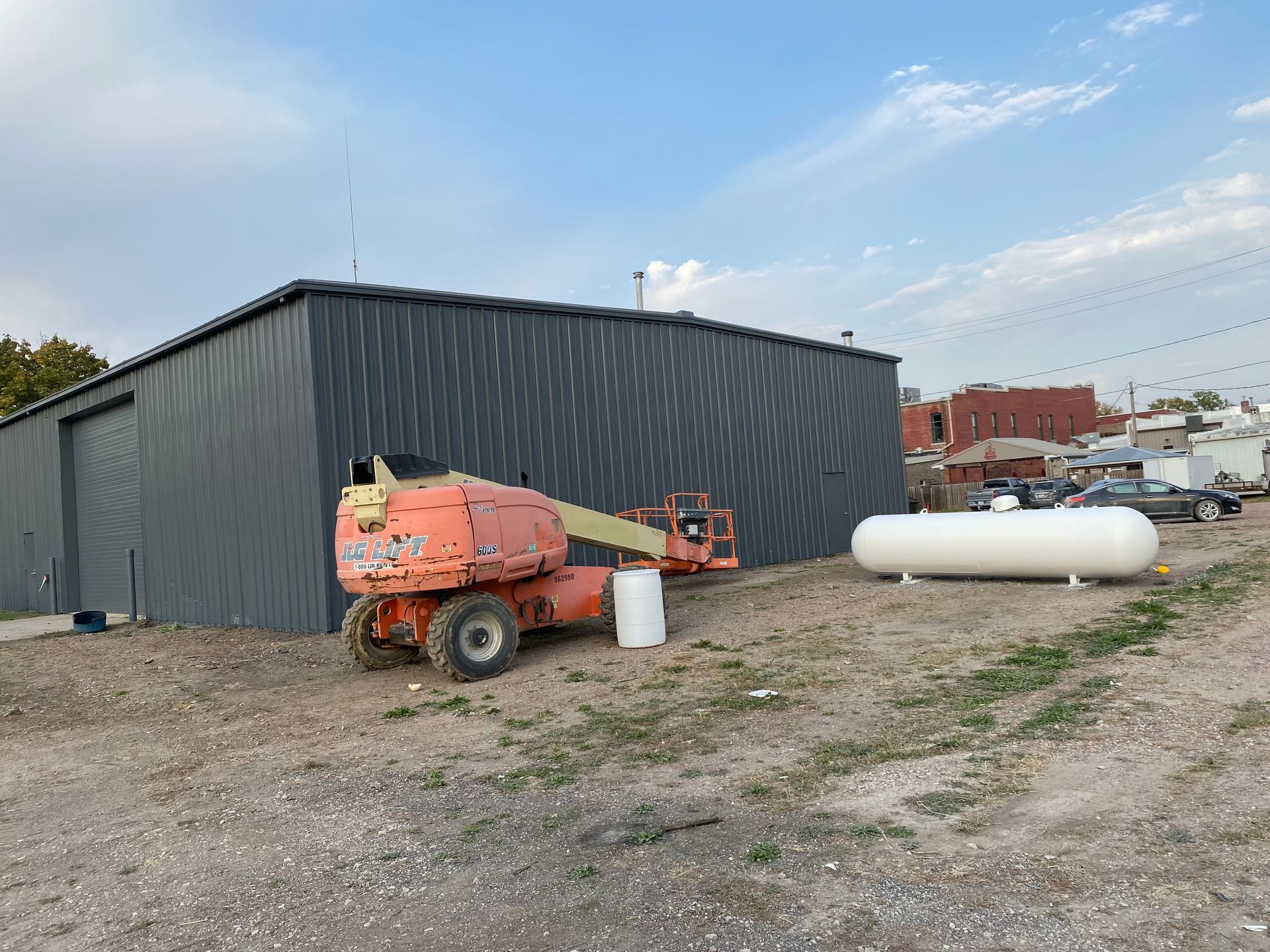 A metal building with a cherry picker, propane tank, and dirt ground. Blue sky with a few clouds.