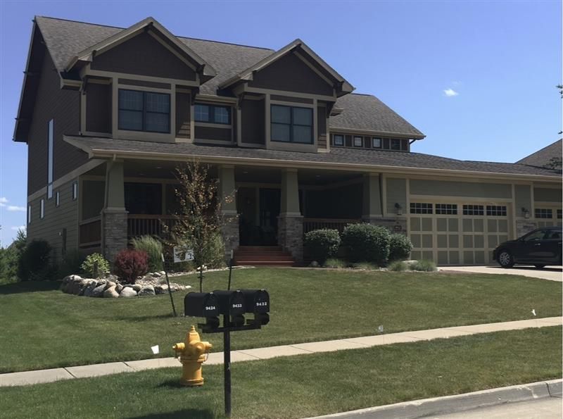 A two-story, dark brown suburban house with a covered front porch, stone pillars, a two-car garage, and a lawn.