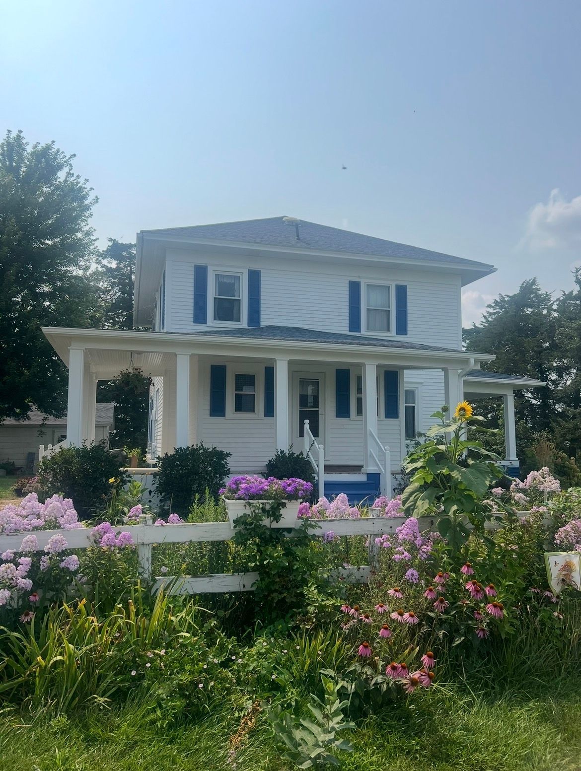 A white two-story house with blue shutters and a front porch, fronted by a lush flower garden with a white fence.
