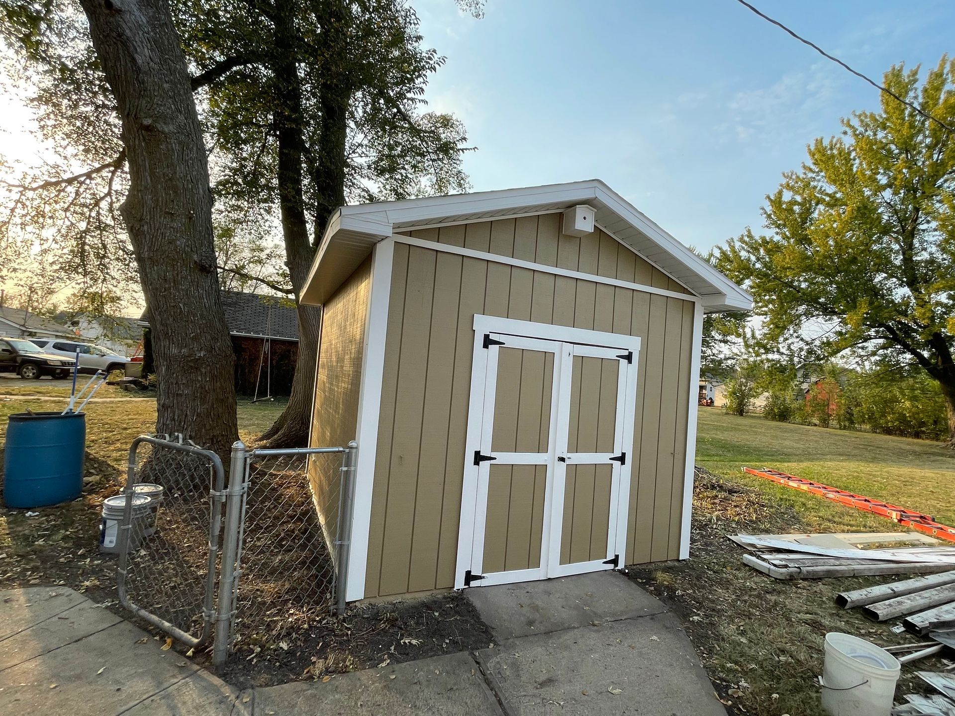 Tan shed with white trim, built on a slope near a tree and chain link fence.