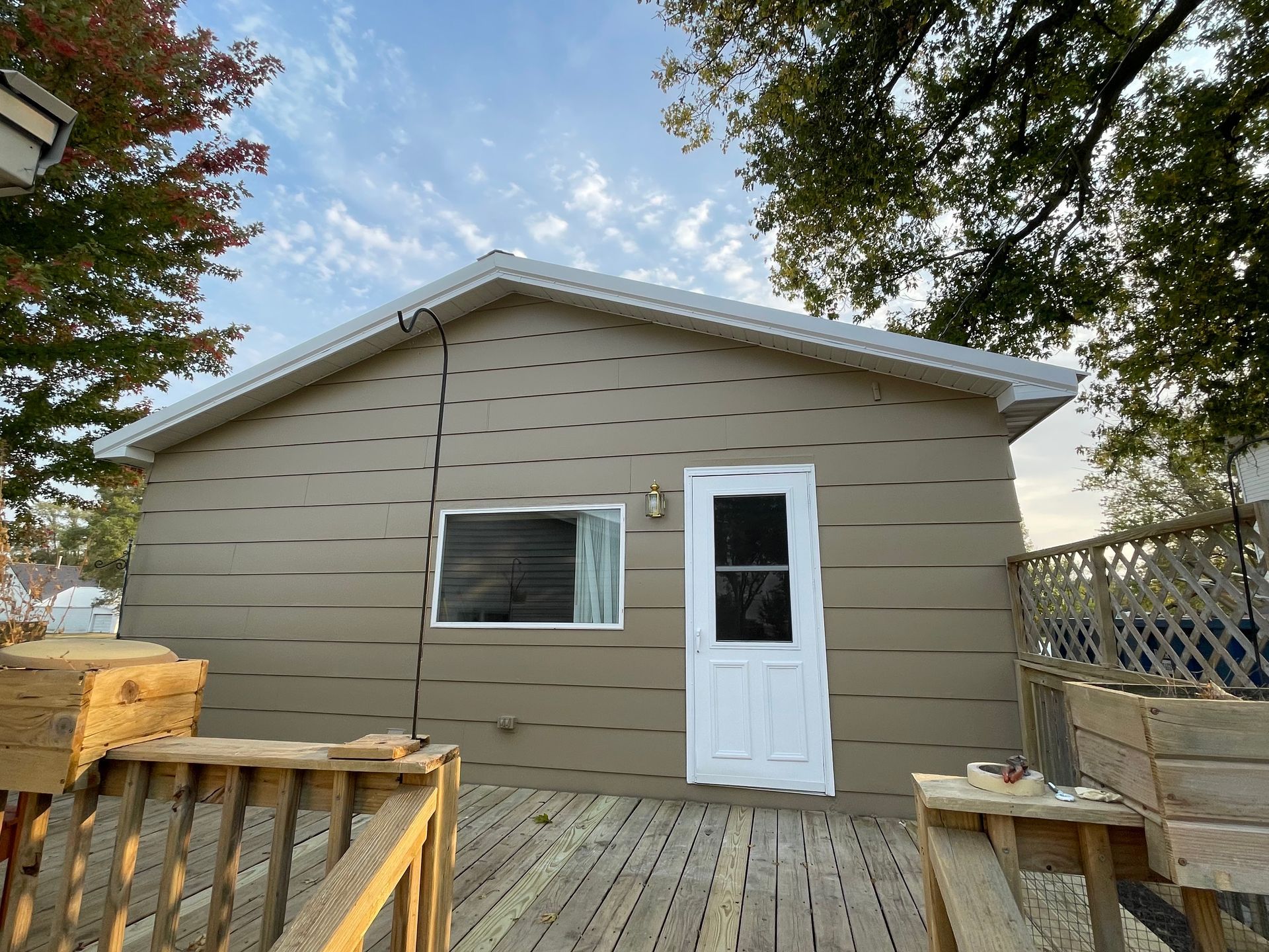 Tan house with white trim, window, and door. Wooden deck in front with fall foliage and cloudy sky.