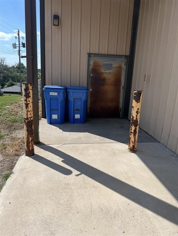 Exterior view of a building side door with two blue trash bins and two rusted metal bollards on a concrete pad.