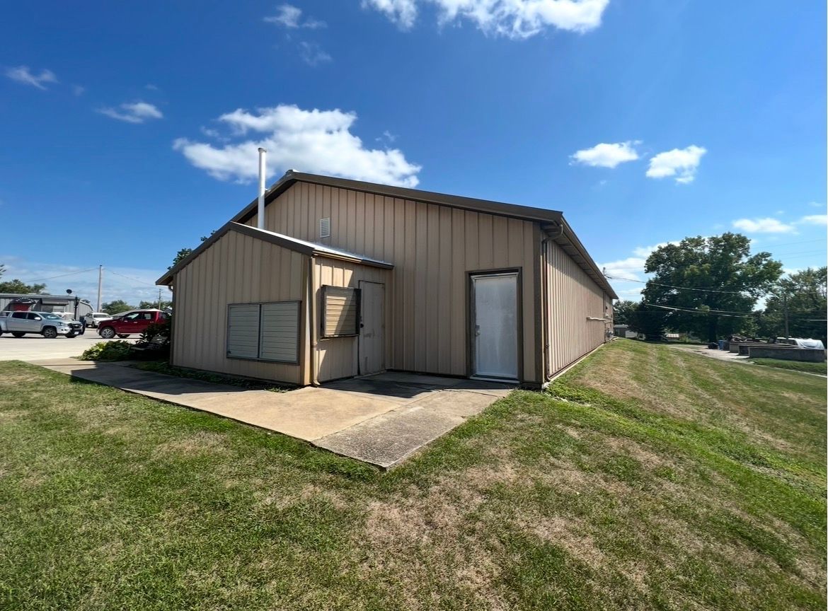 A beige, metal-sided building with a small side addition, a single white door, and a grass lawn under a blue, cloudy sky.