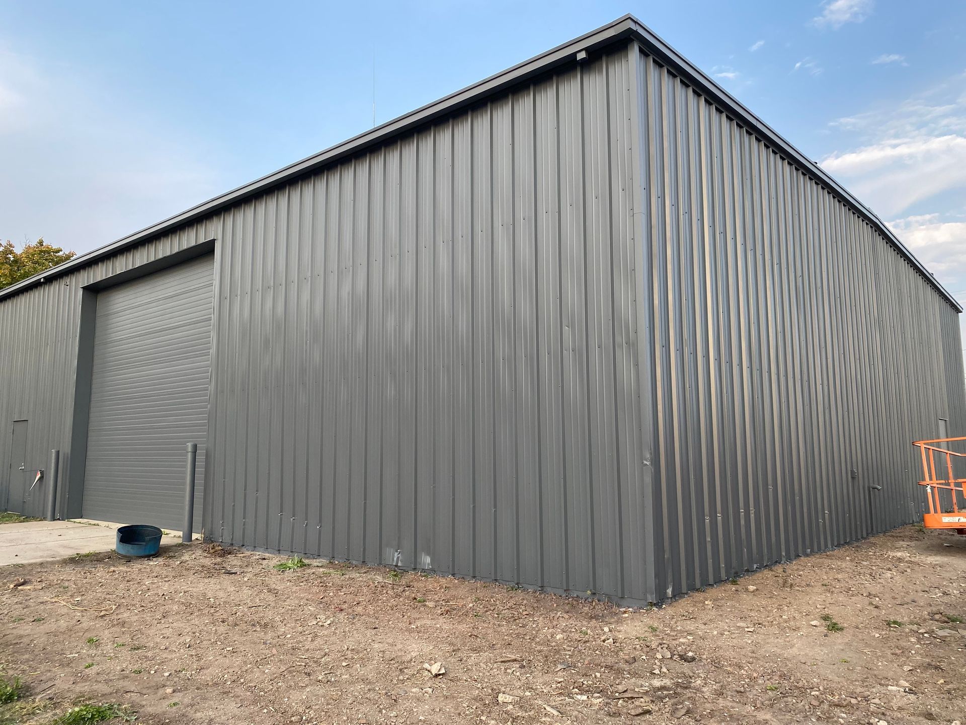 Grey metal building with large sliding door against a blue sky, on a dirt lot.