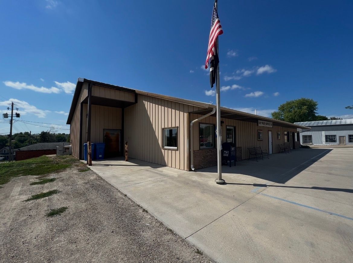 A beige, single-story metal building with a parking lot and an American flag on a pole under a clear blue sky.