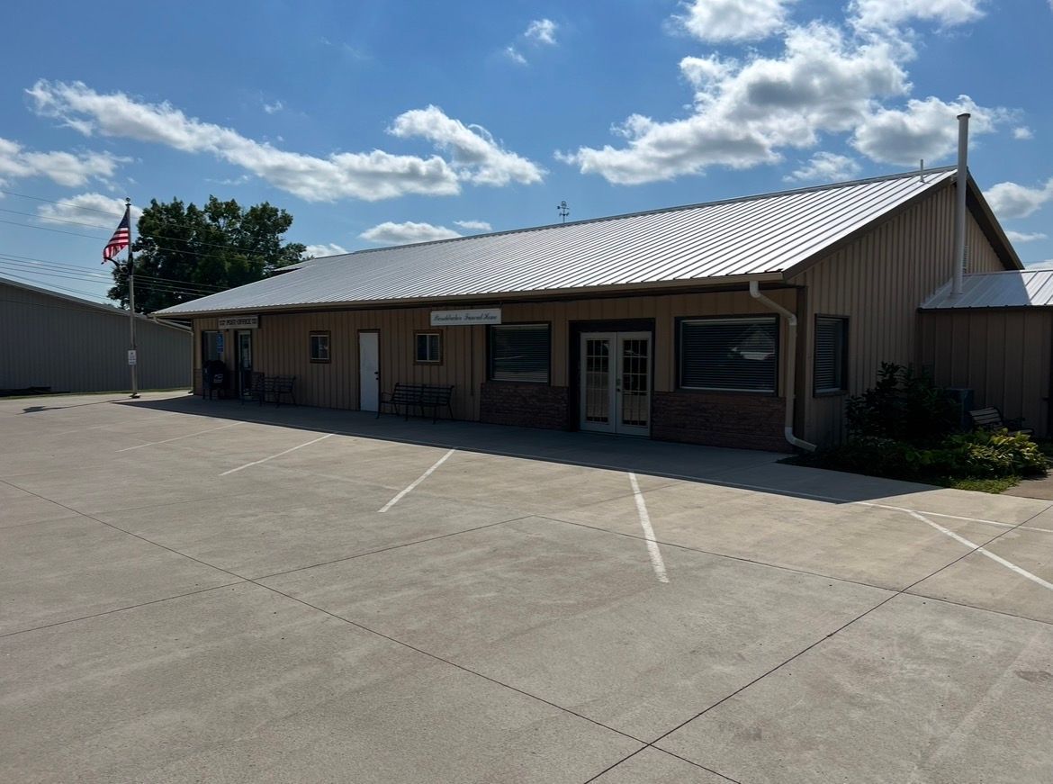 A tan, single-story commercial building with a metal roof and a large concrete parking lot under a blue sky.