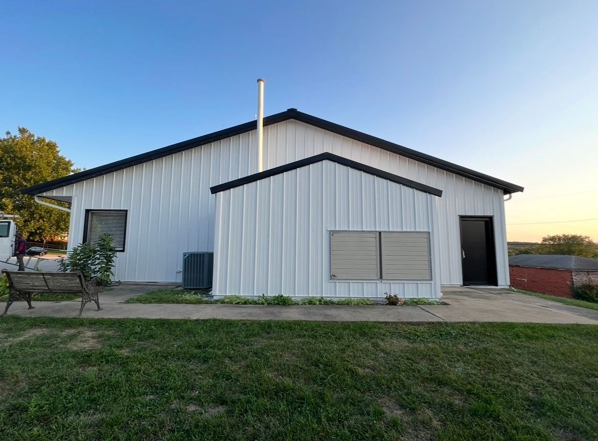 A white, metal-sided building with a dark roof and chimney, featuring a door, vents, and a bench on a concrete patio.