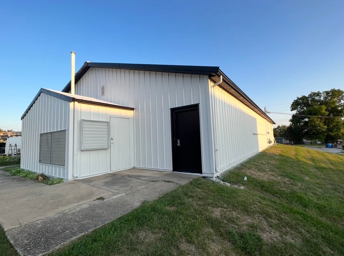 A white, metal-sided building with a black door and a small side extension under a clear, blue sky.