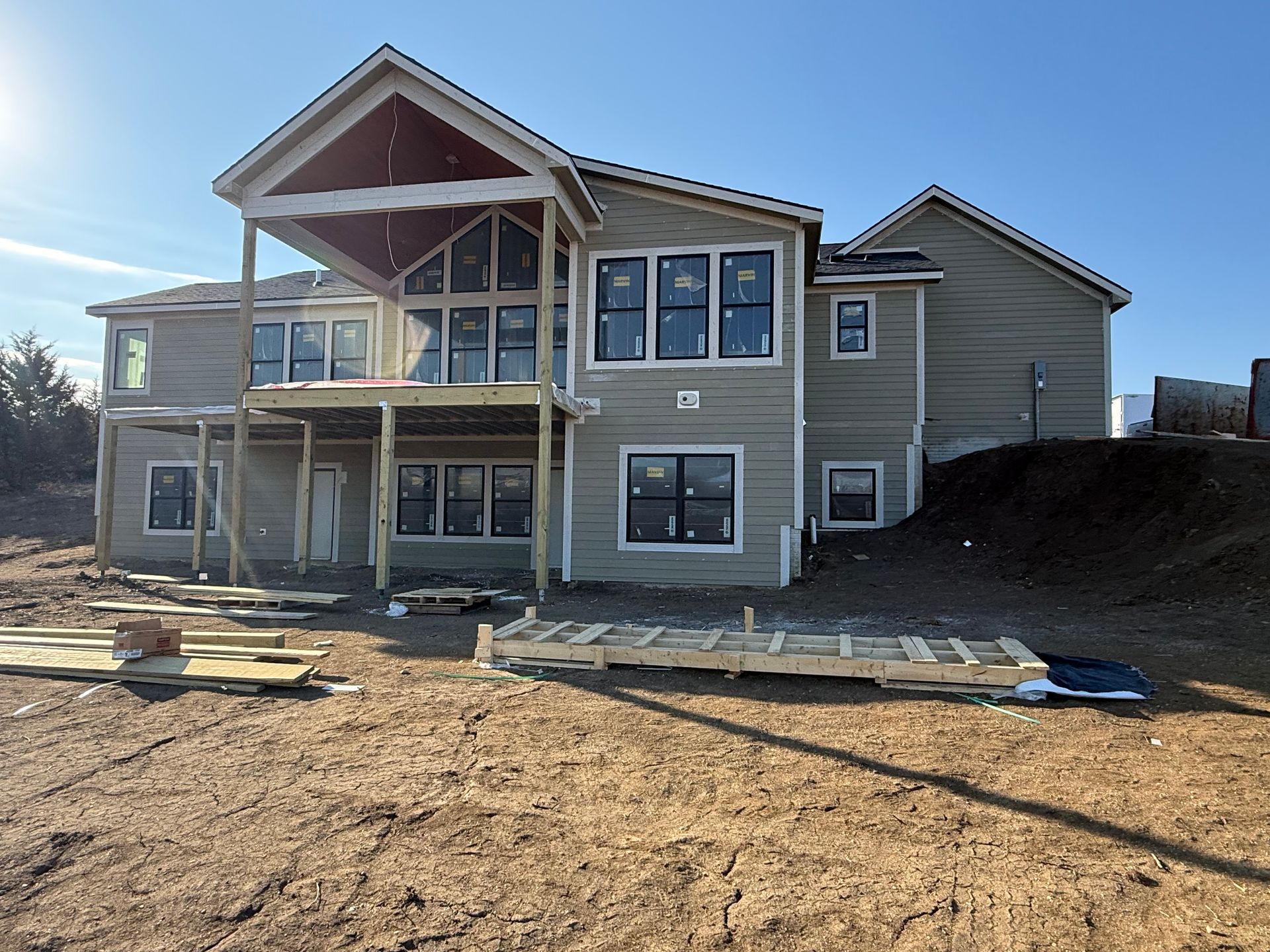 Two-story house under construction with light-colored siding, black-framed windows, and a partially built porch. Sunny day, exterior shot.