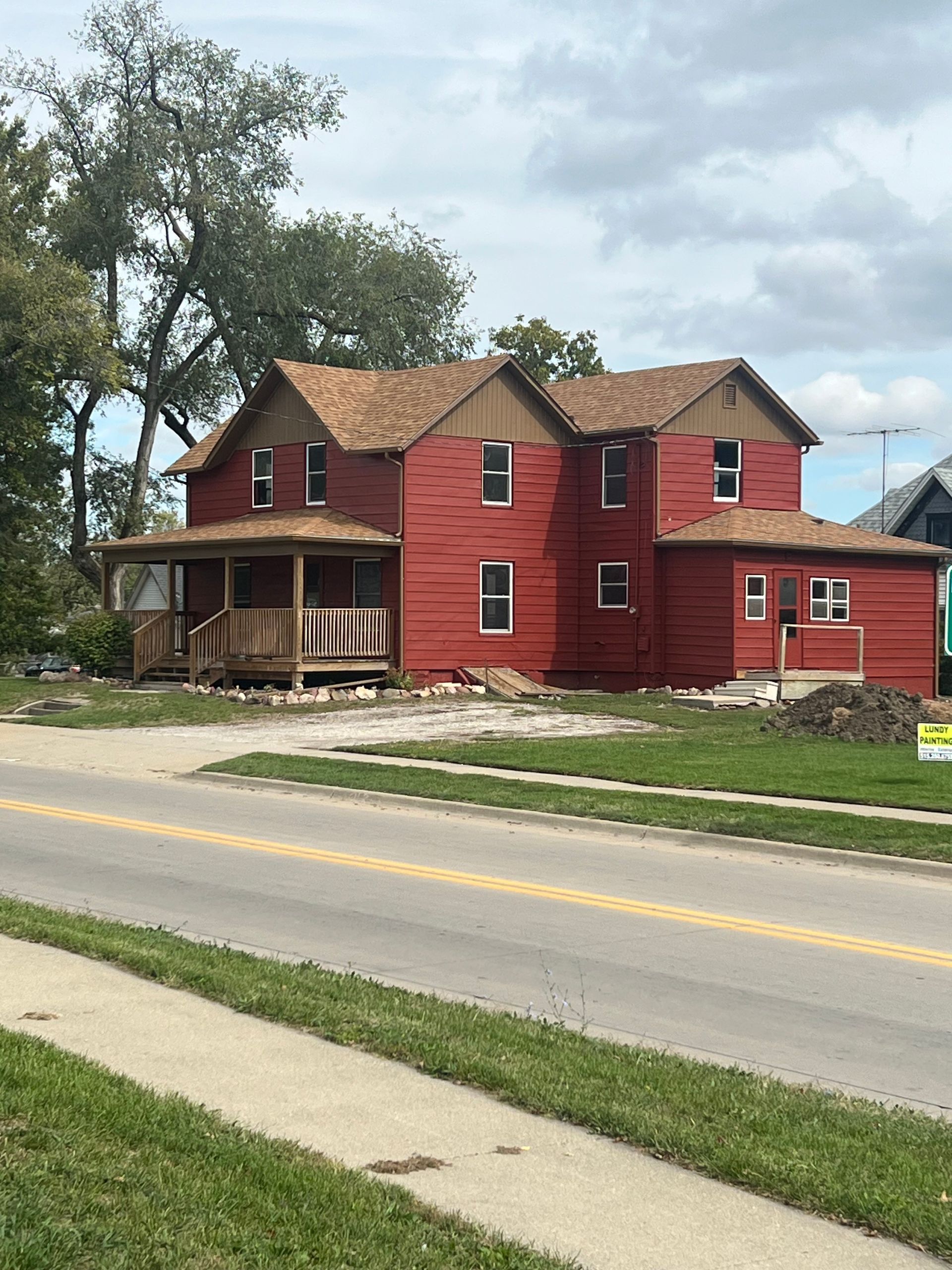 Red two-story house with a wrap-around porch, brown roof, and roadside.