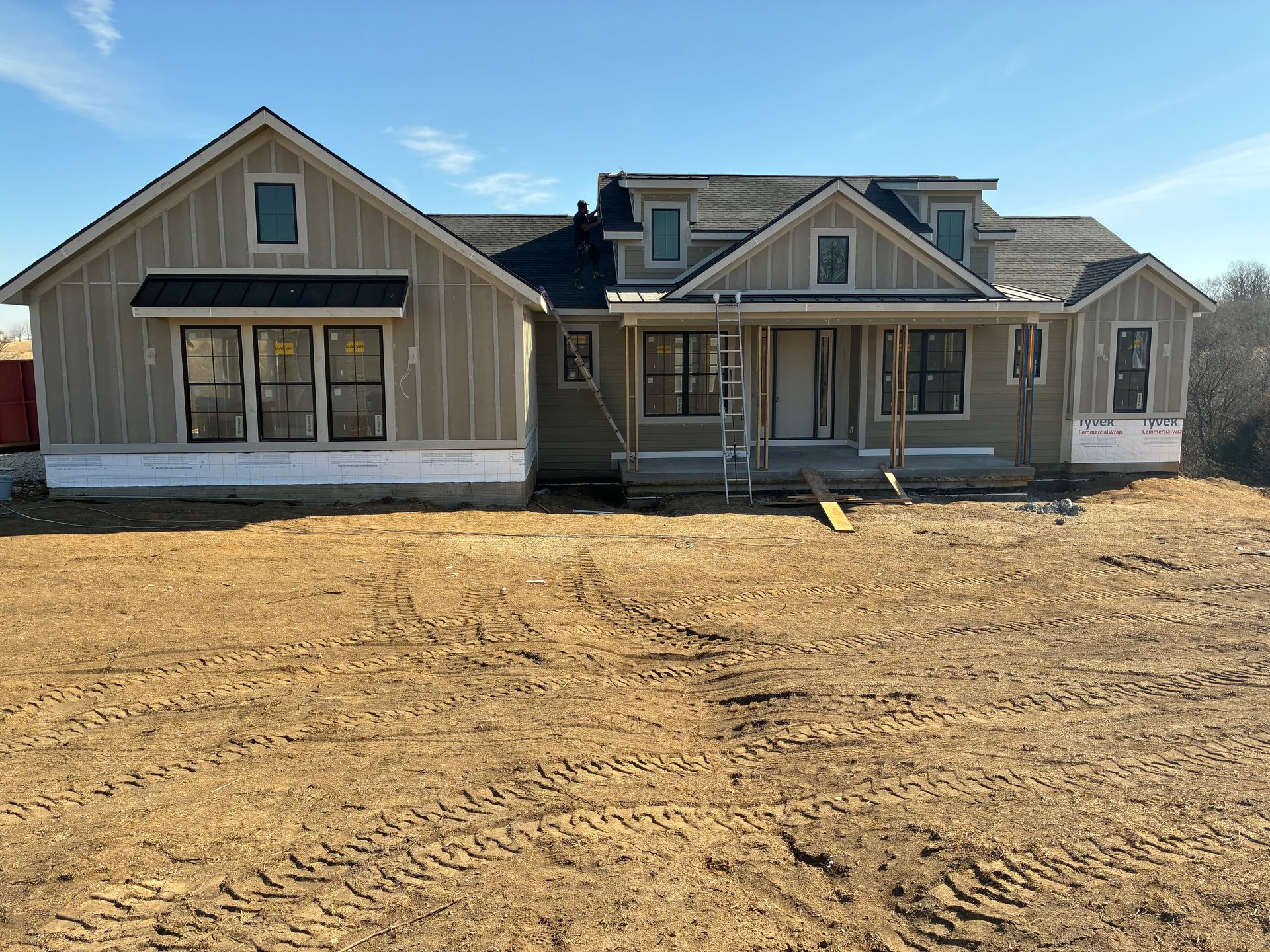 New house under construction with light siding and black roof on a dirt lot, clear sky.