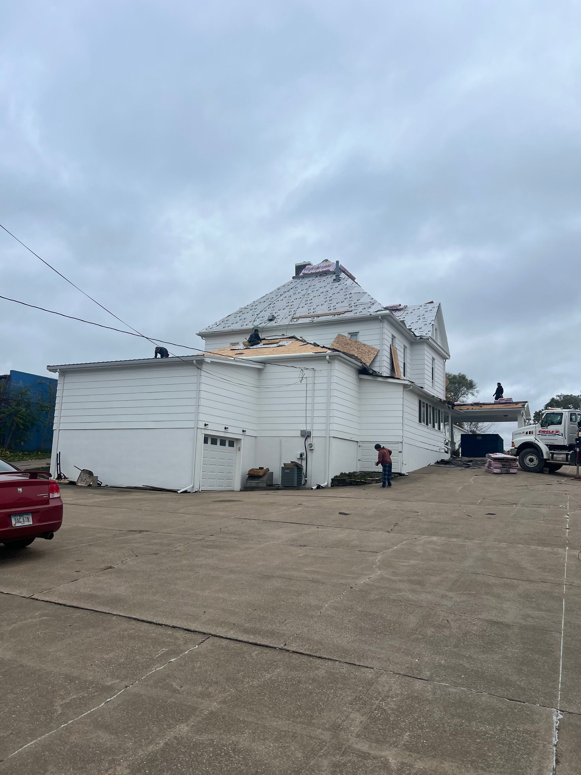 White building with roof repair in progress; workers on roof and ground. Trucks and cloudy sky.