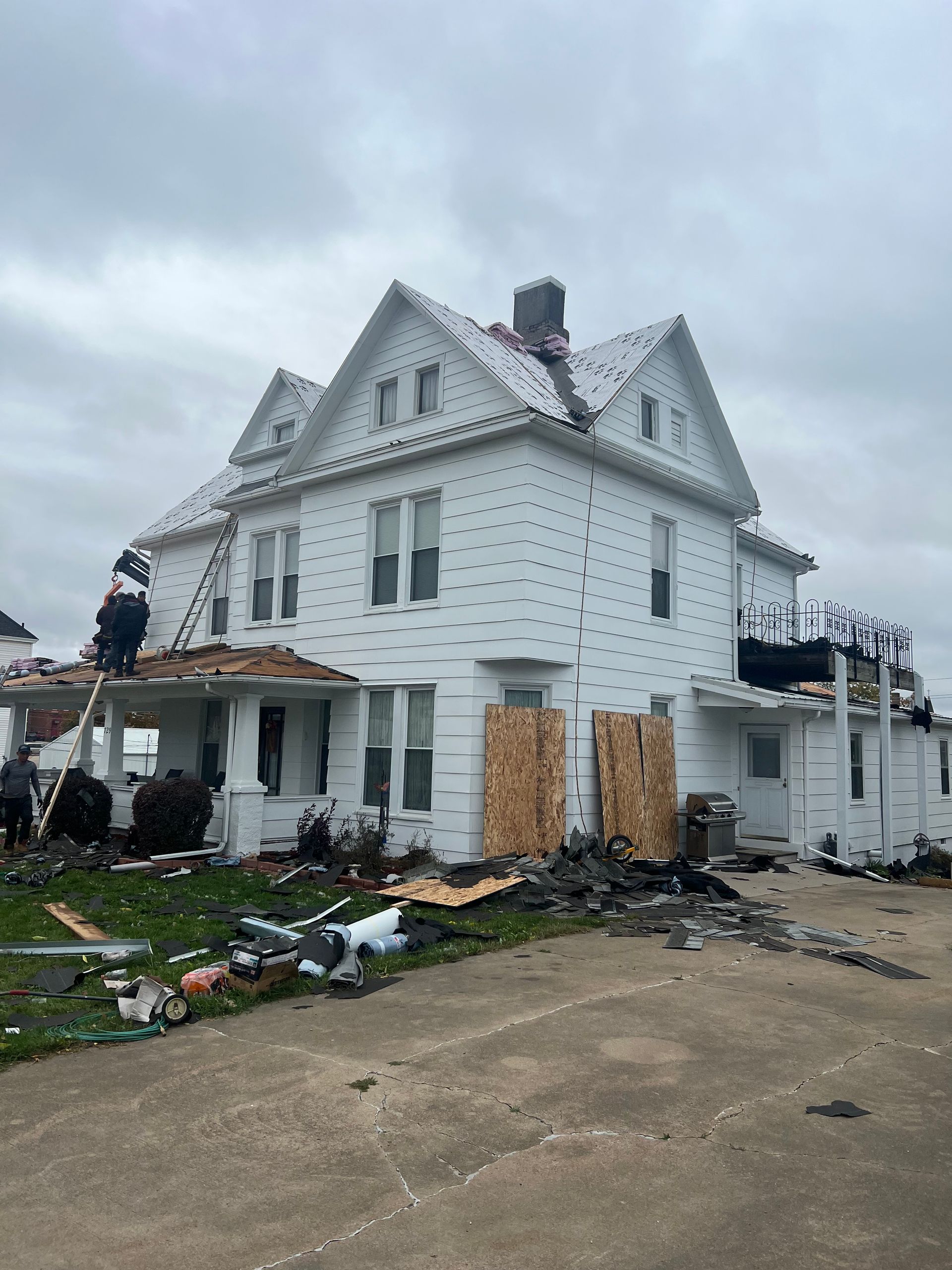 White house with roof damage and workers on a ladder, debris scattered around, cloudy sky.