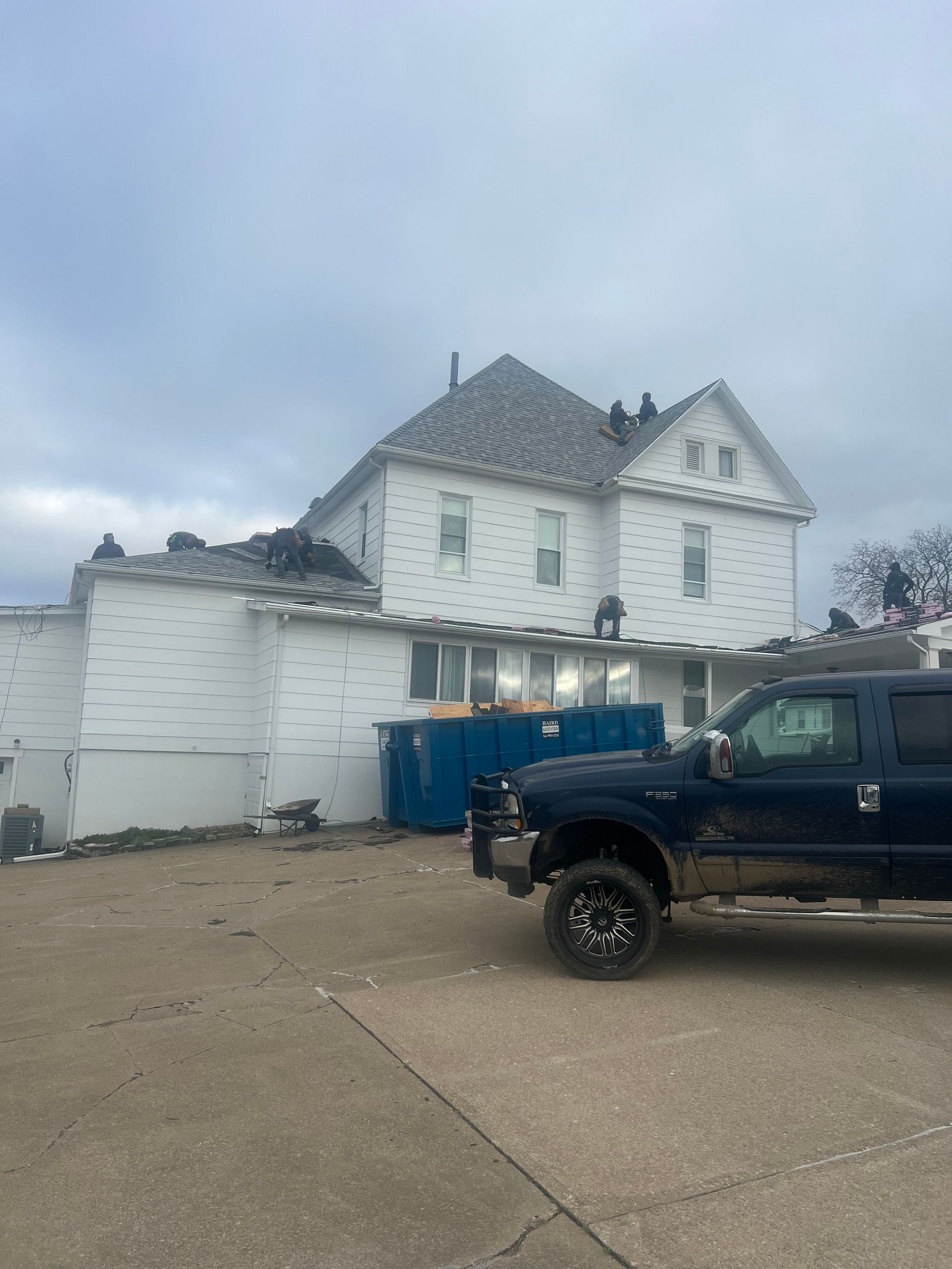 Workers on roof of white building, blue dumpster, and dark pickup truck. Cloudy sky.