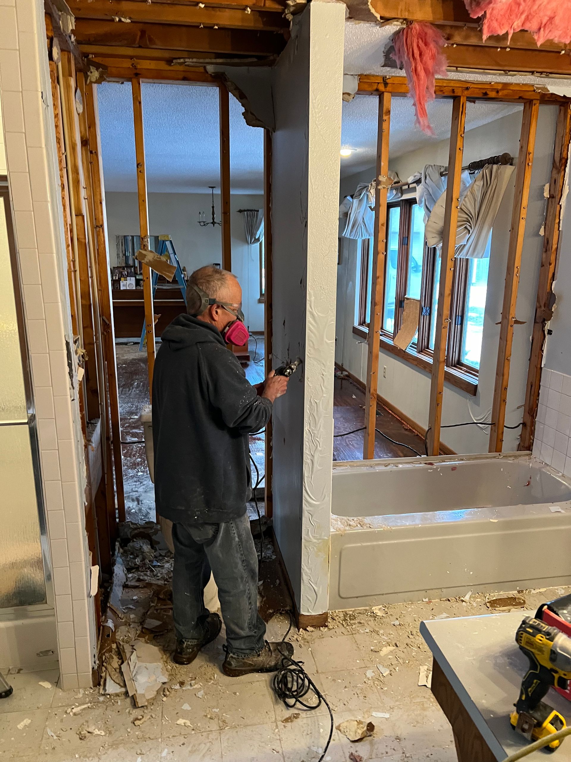 Man in a respirator is working on bathroom demolition, with exposed framing, debris, and a partially removed tub.