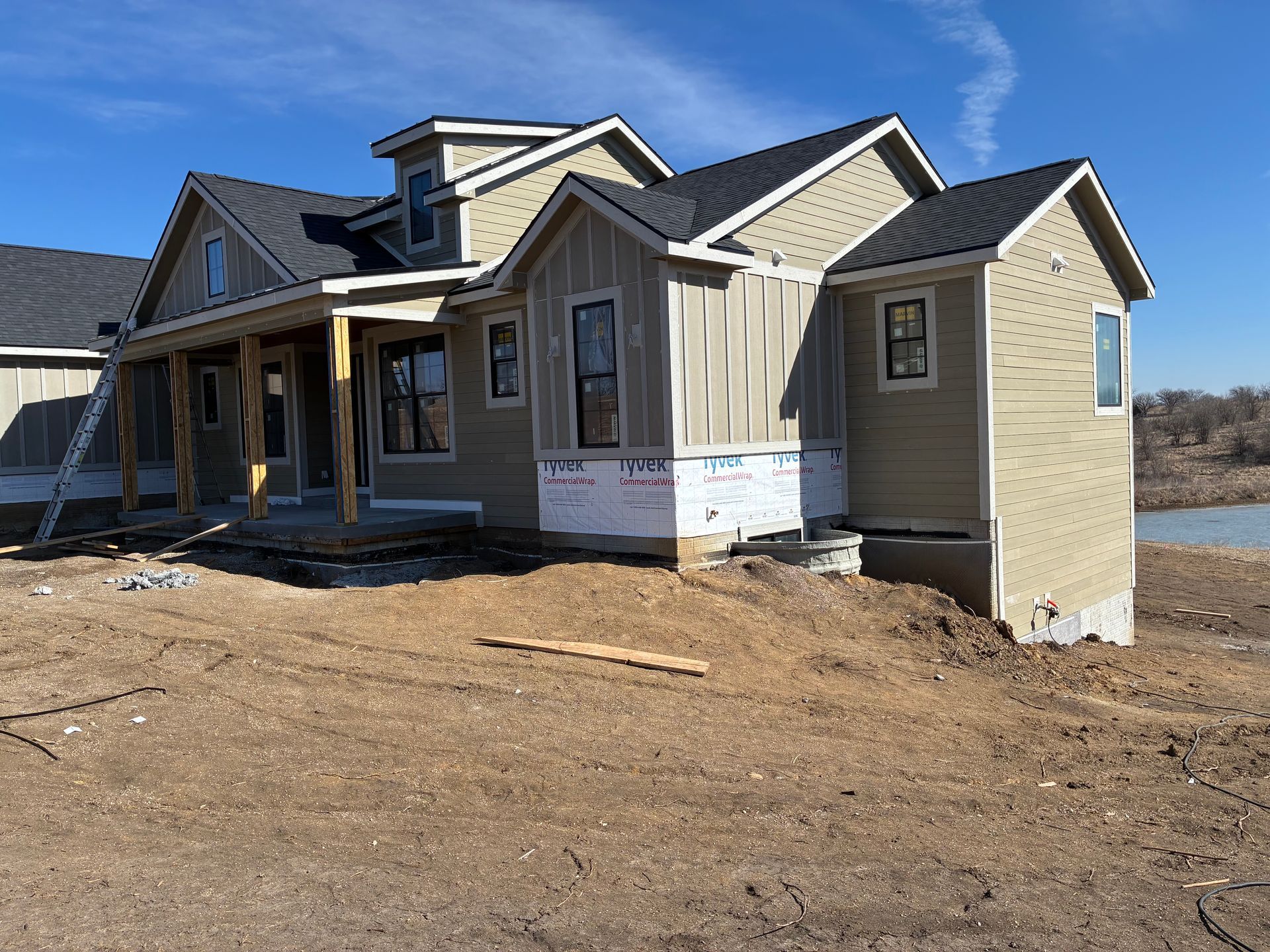 New house under construction with light brown siding, black roof, and dirt ground.