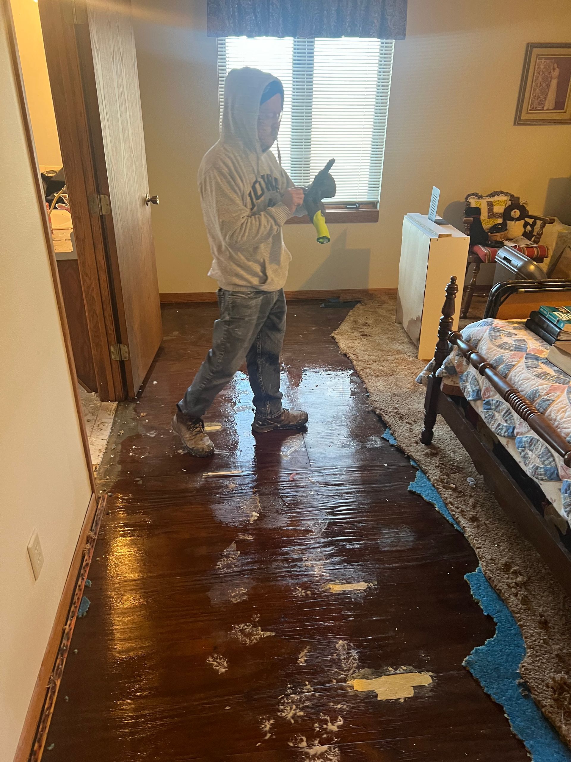 Man in a flooded room, pointing, wearing a hoodie. Water covers the wood floor, near a window and bed.