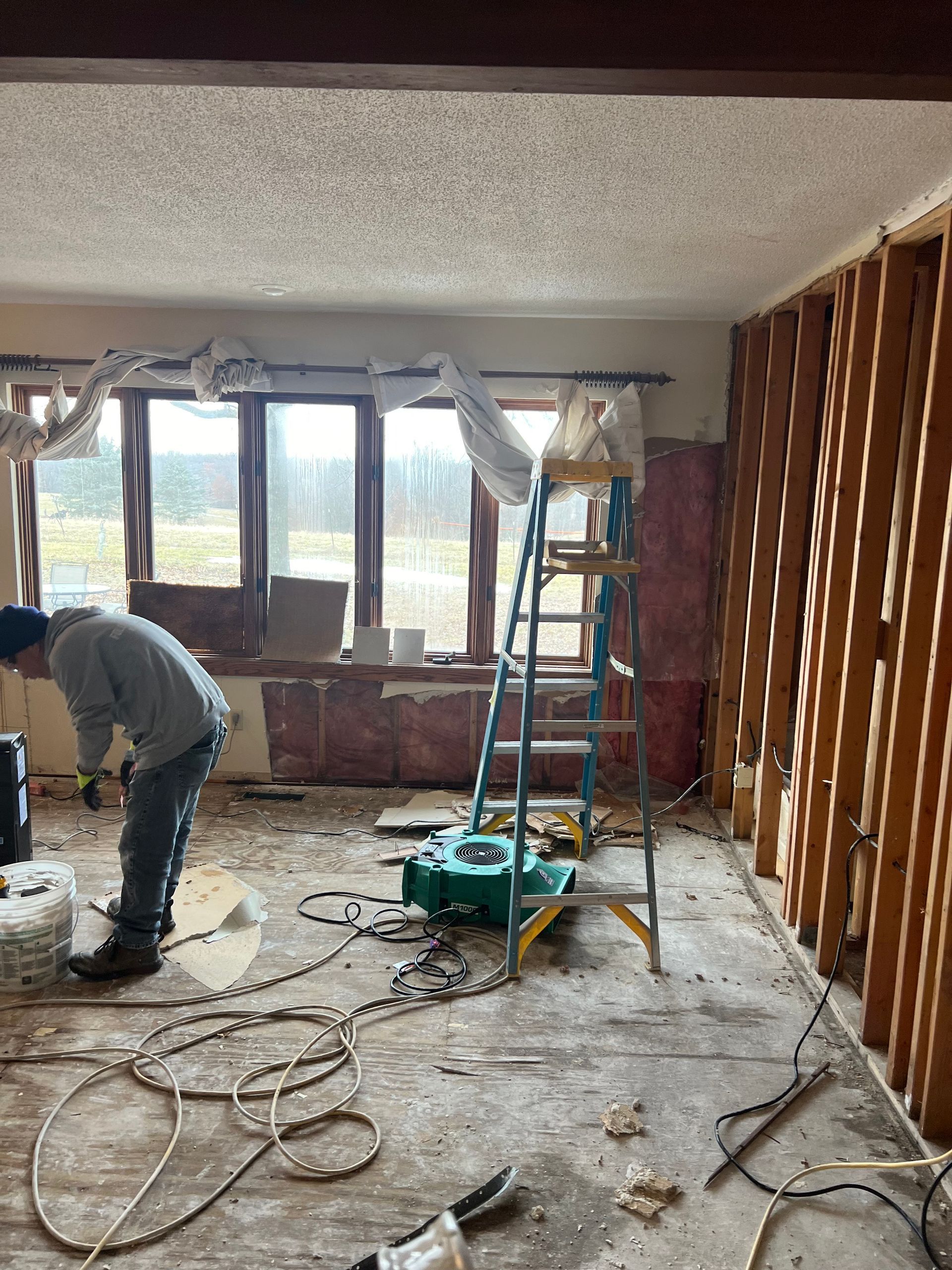 Man working in room undergoing renovation, with exposed framing, ladder, and equipment.