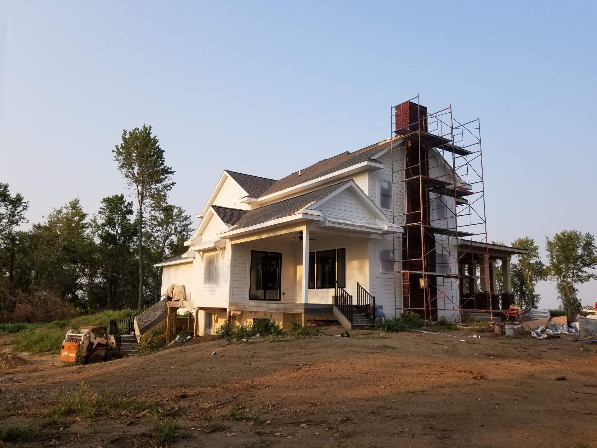 House under construction with scaffolding around the chimney and exposed siding, on a brown, dirt lot.