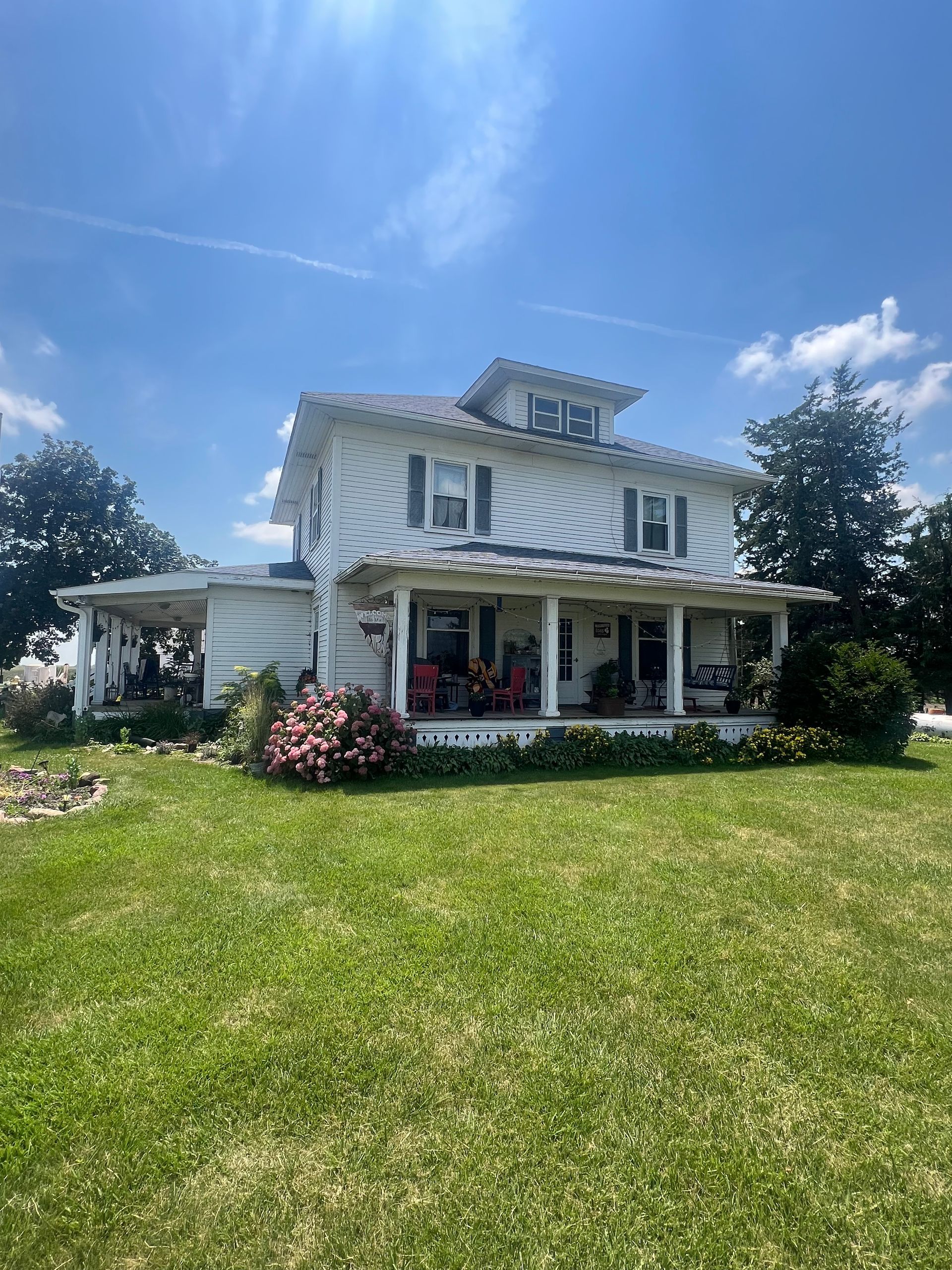 White farmhouse with wrap-around porch, blue shutters, and green lawn under a blue sky.