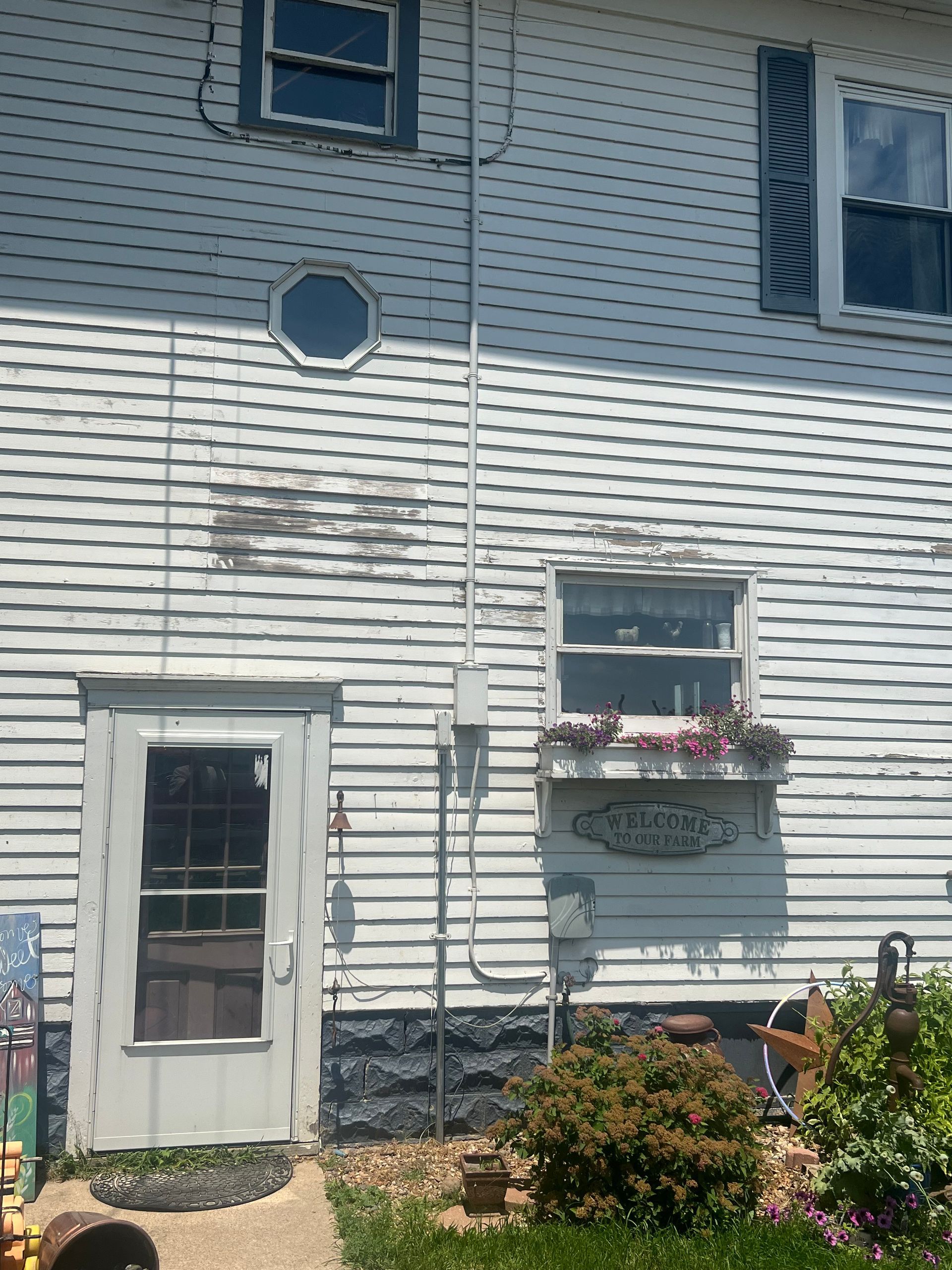White clapboard house with weathered paint, multiple windows, a door, and some plants.