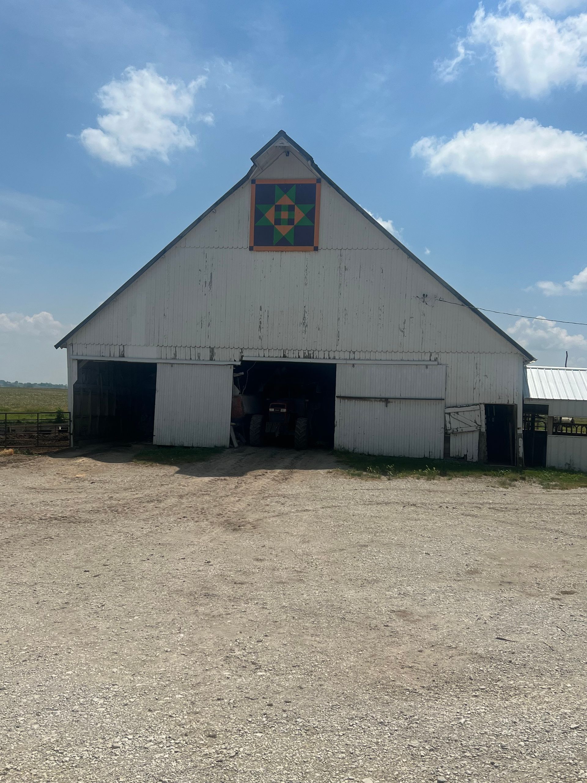 White barn with quilt square on gable, on a gravel lot under a partly cloudy sky.