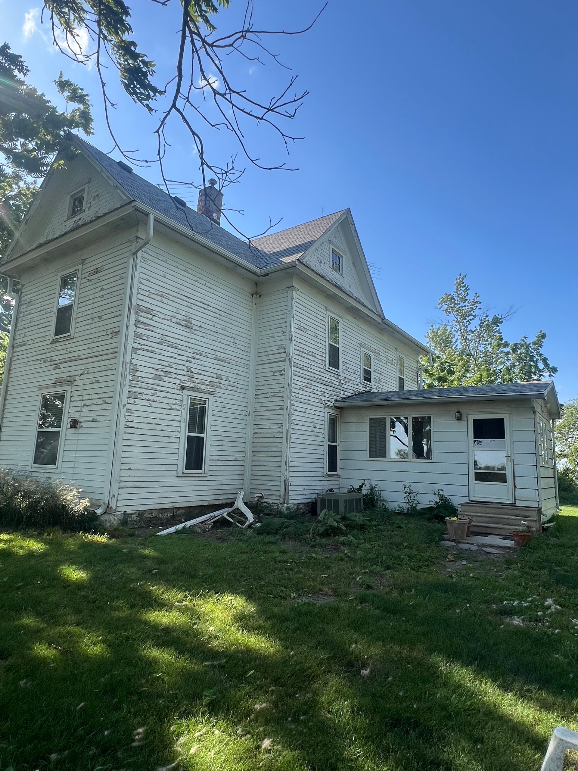 White two-story house with peeling paint and an attached small side room, set against a blue sky.