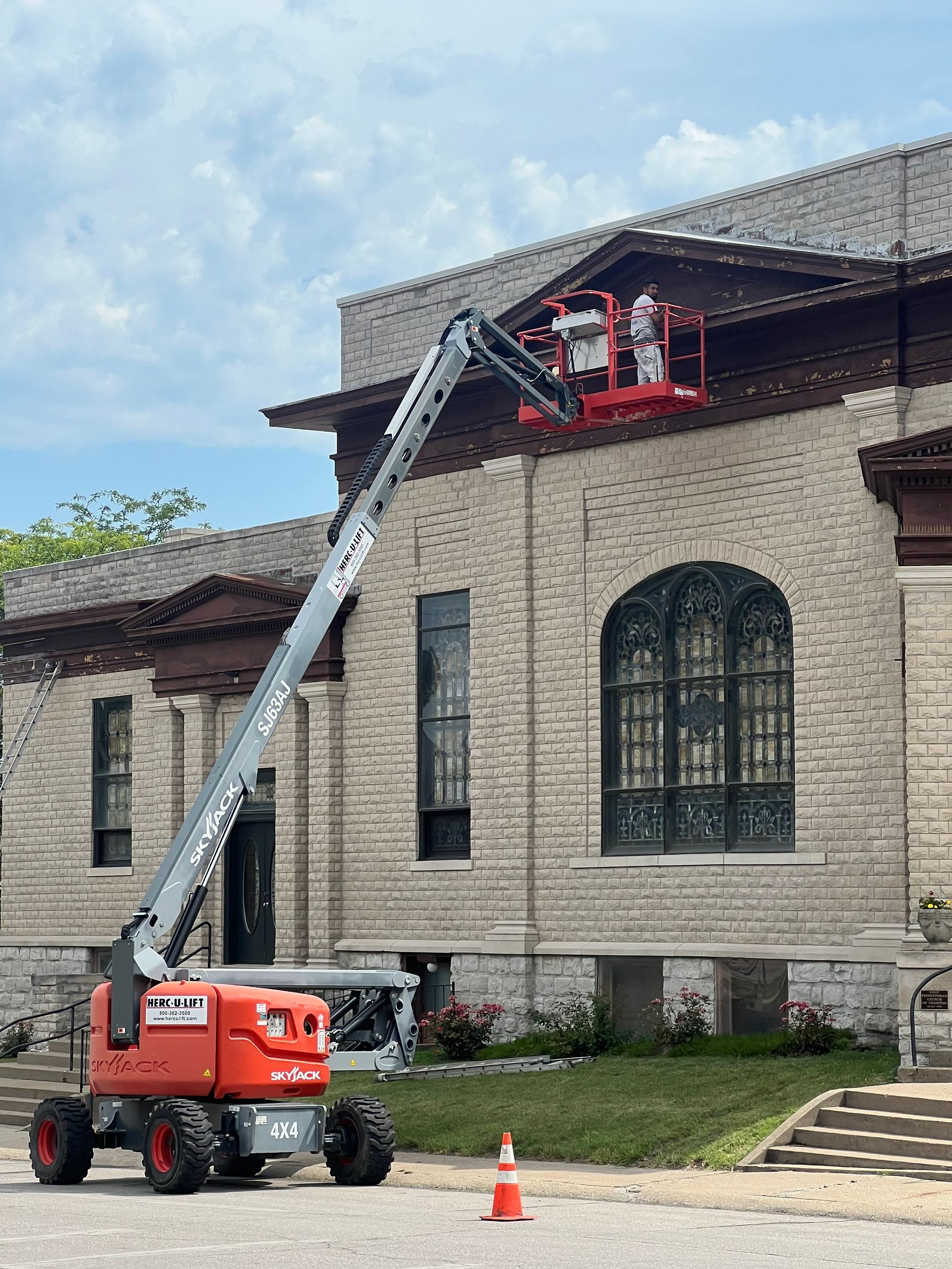An orange lift machine is raised to the roof of a brick and stone building. A person is in the lift basket.