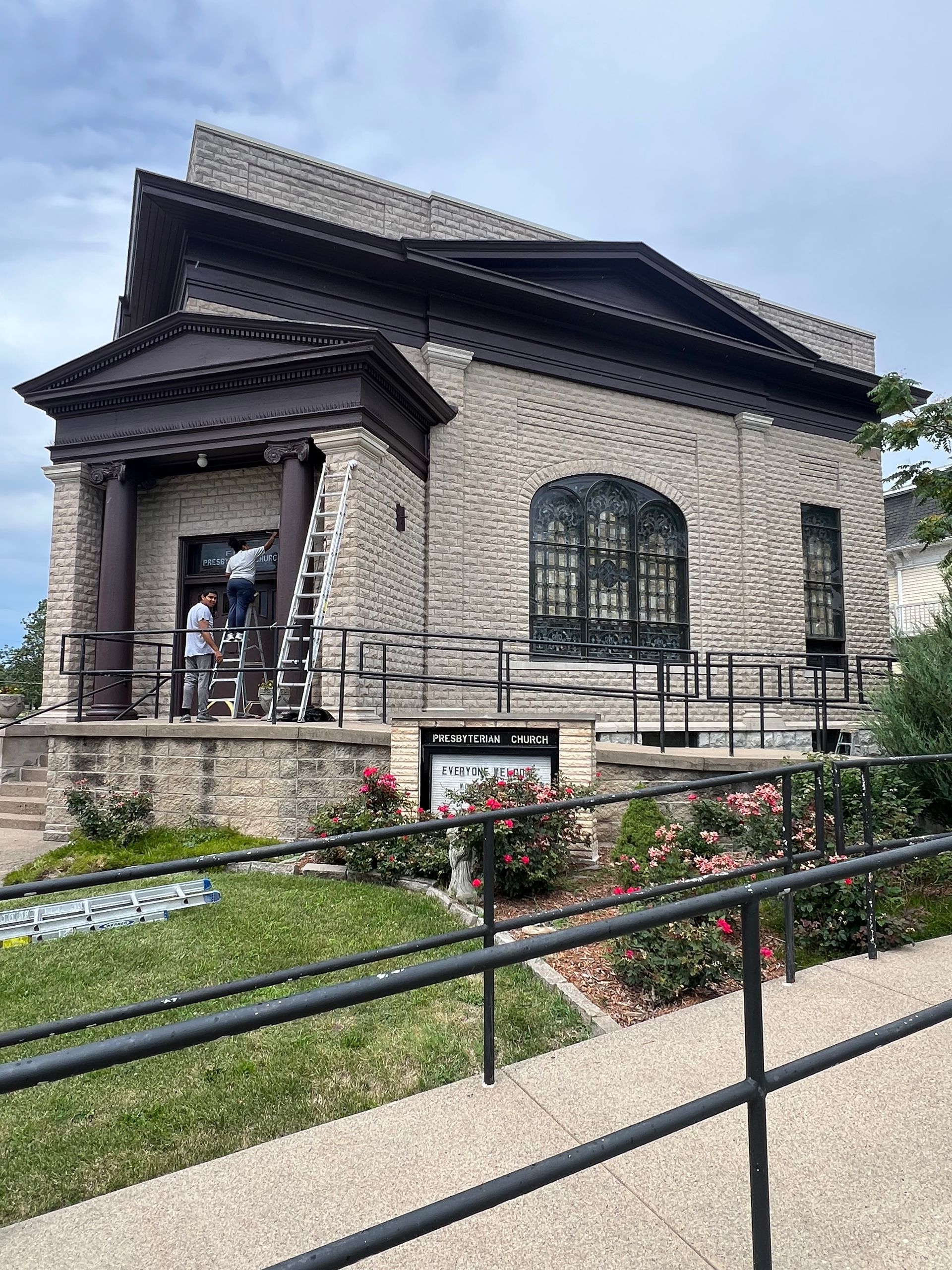 Stone building with brown trim. Two people on a ladder near the entrance. Ramp and rose bushes in front.