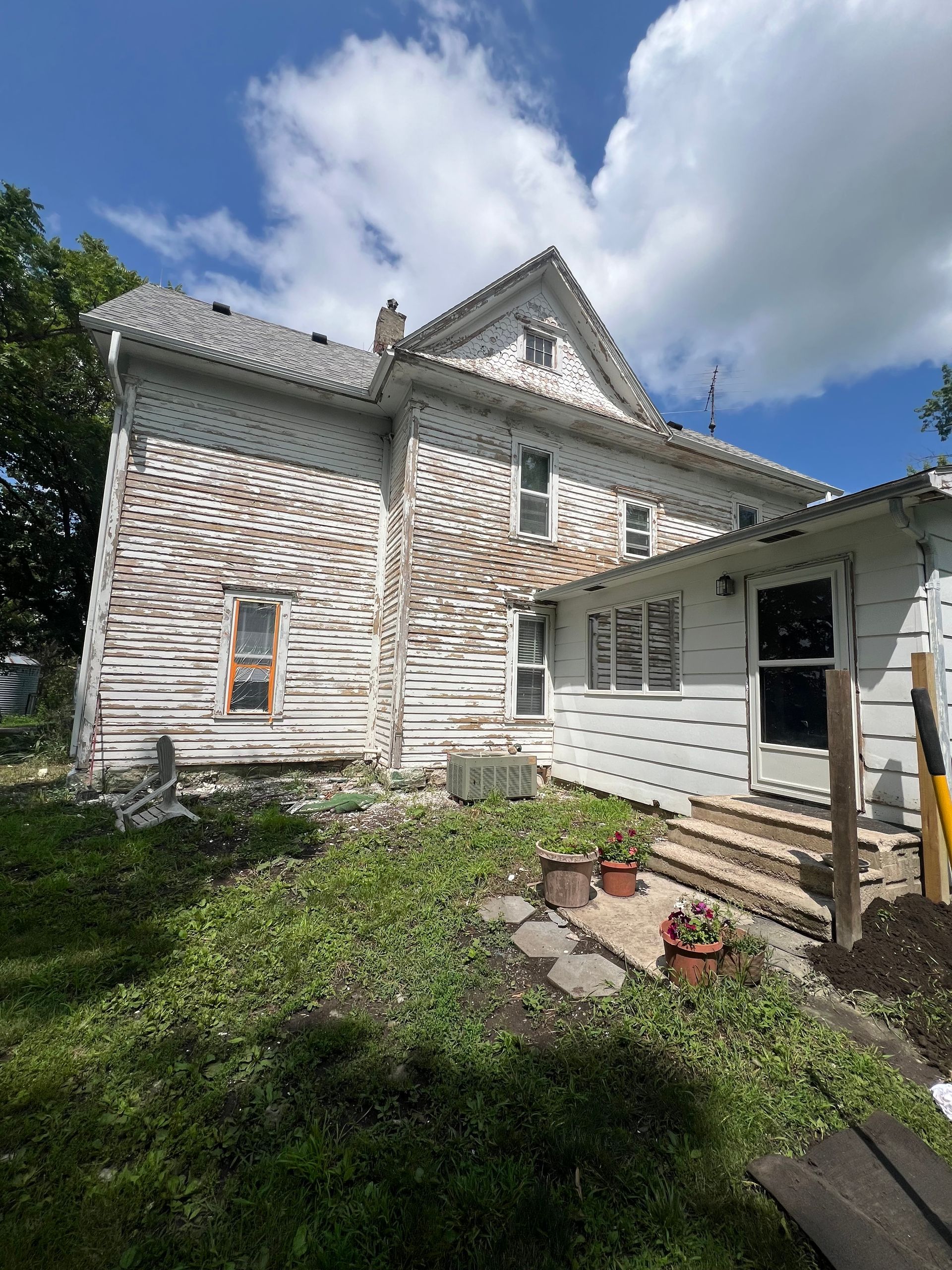 A weathered, white two-story house with peeling paint and overgrown yard under a partly cloudy sky.