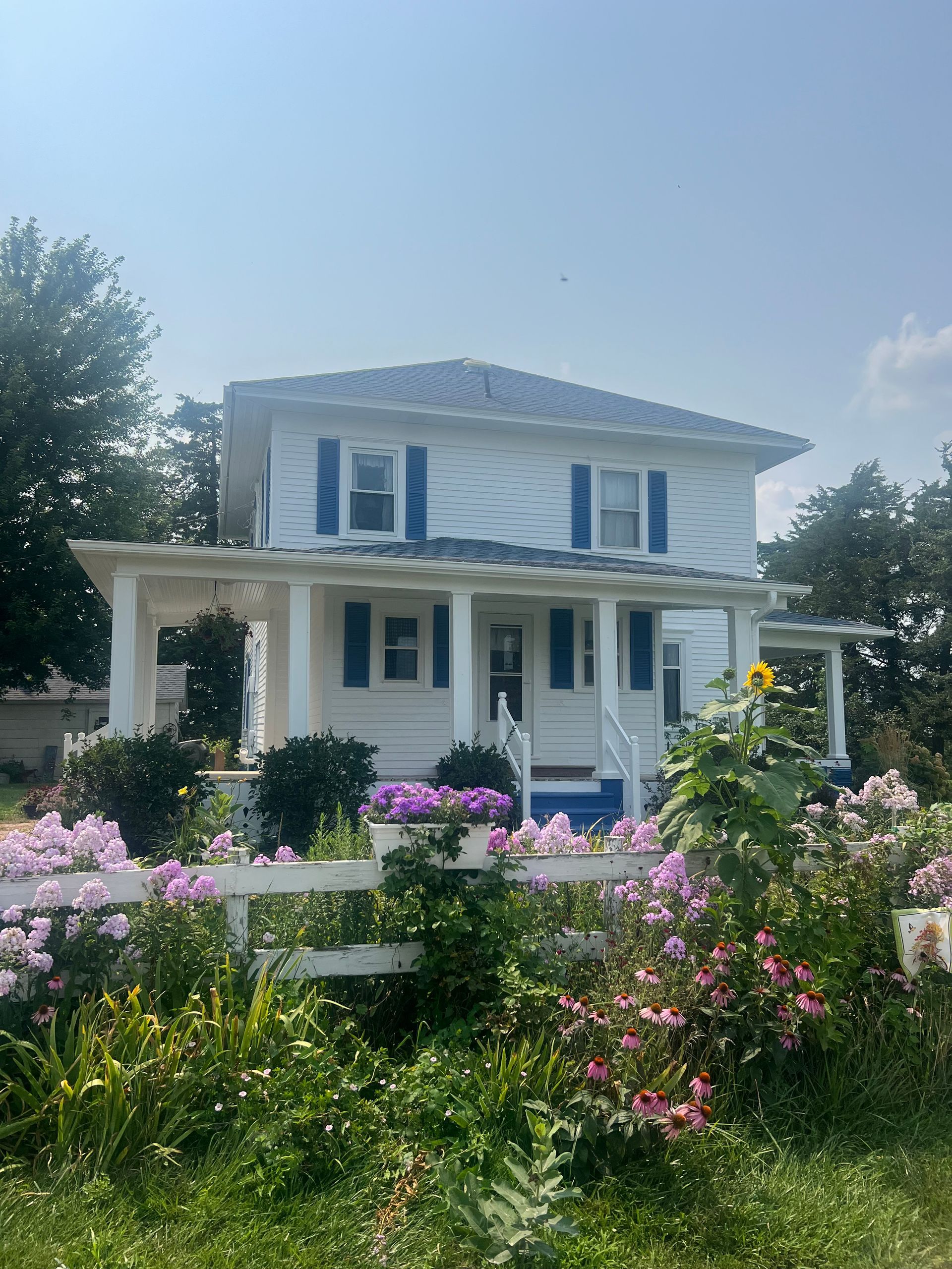 White house with blue shutters, covered porch, and a colorful flower garden.