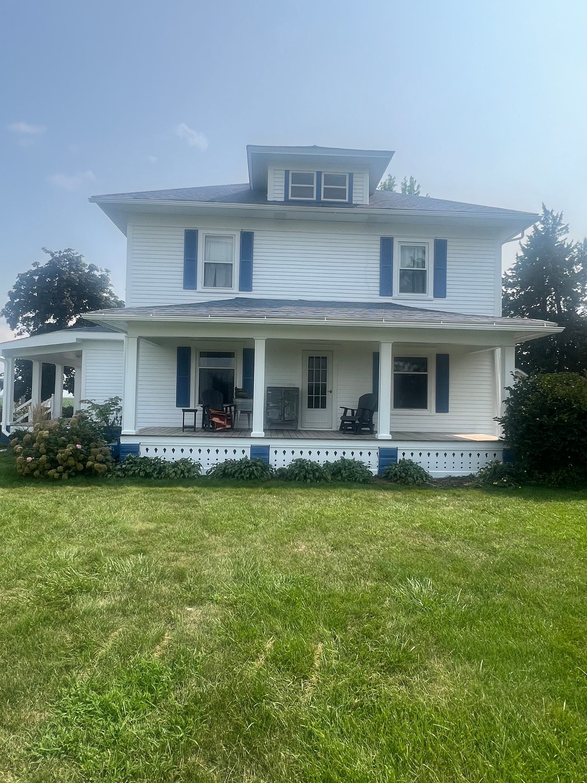 Two-story white house with blue shutters and a porch under a blue sky. Green grass in the foreground.