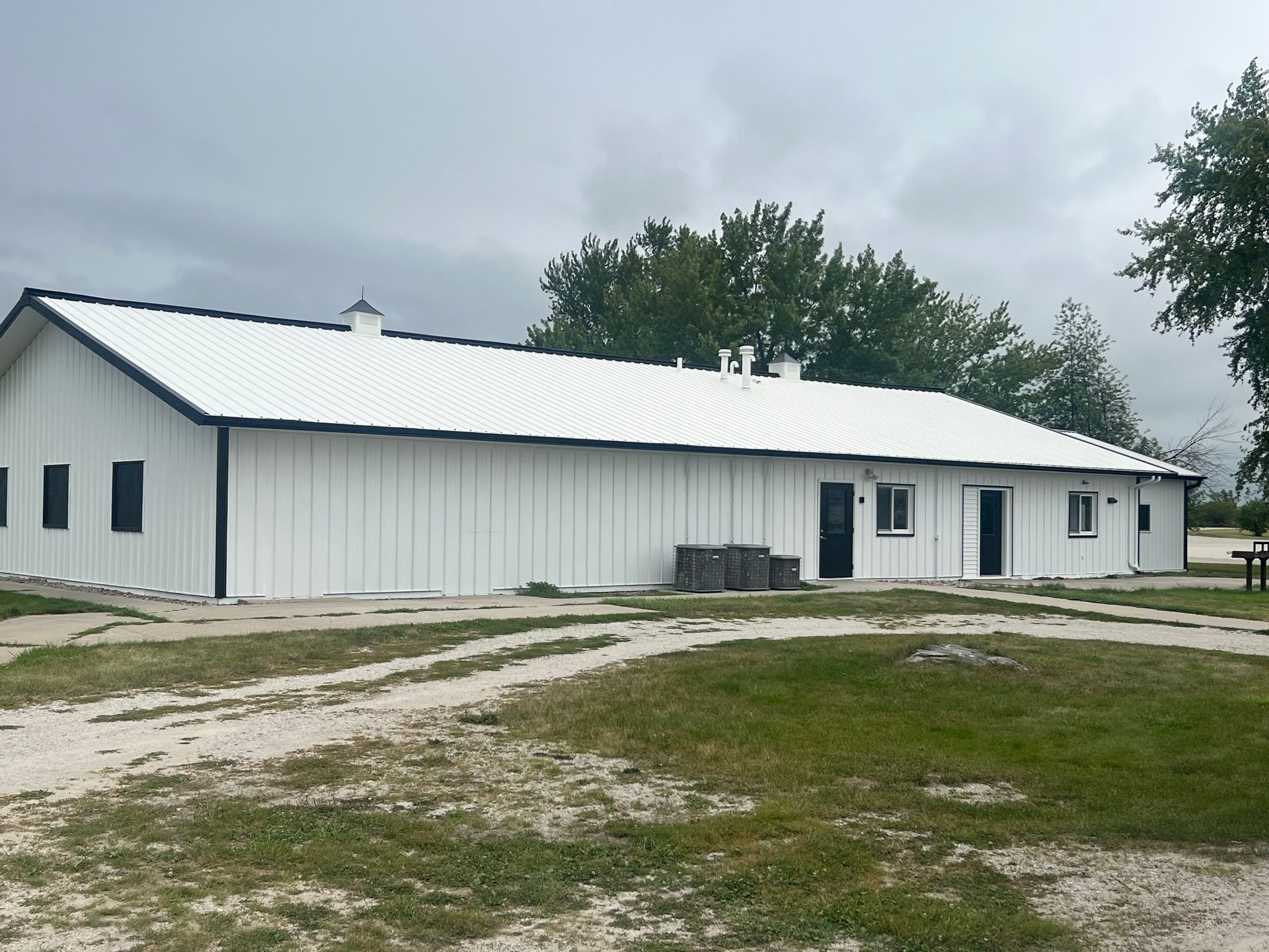 White barn with black trim, a white corrugated metal roof, on a gravel drive, cloudy sky.