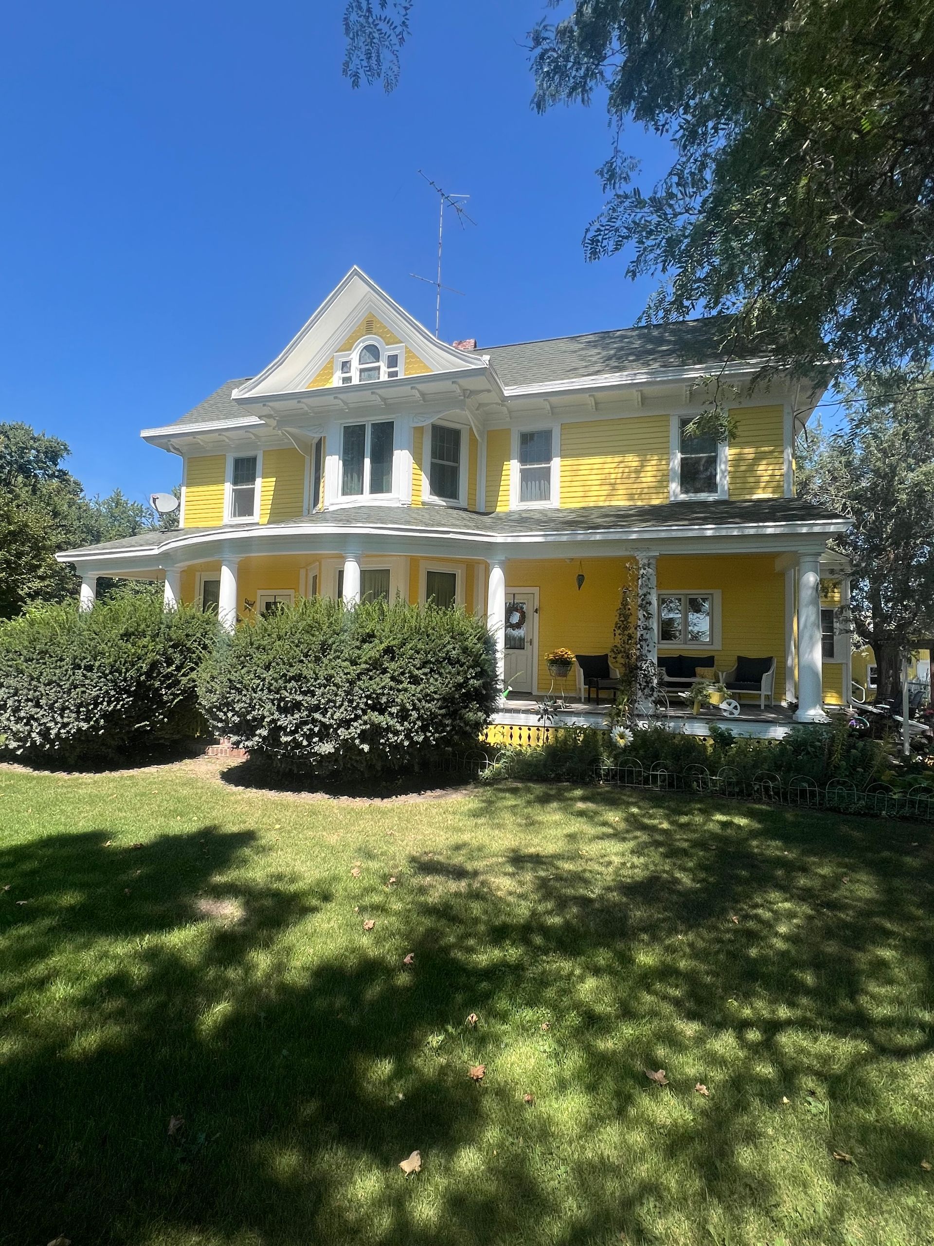 Yellow two-story house with porch. Bushes in front. Sunny day.