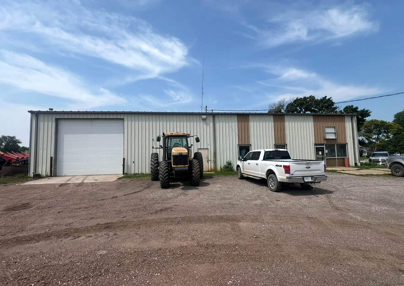 A tractor and white pickup truck parked in front of a metal warehouse under a partly cloudy sky.