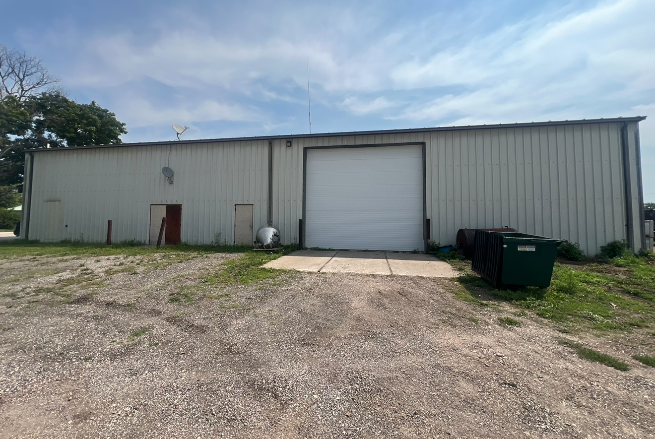 A single-story metal building with a large garage door, gravel lot, and two green trash bins.