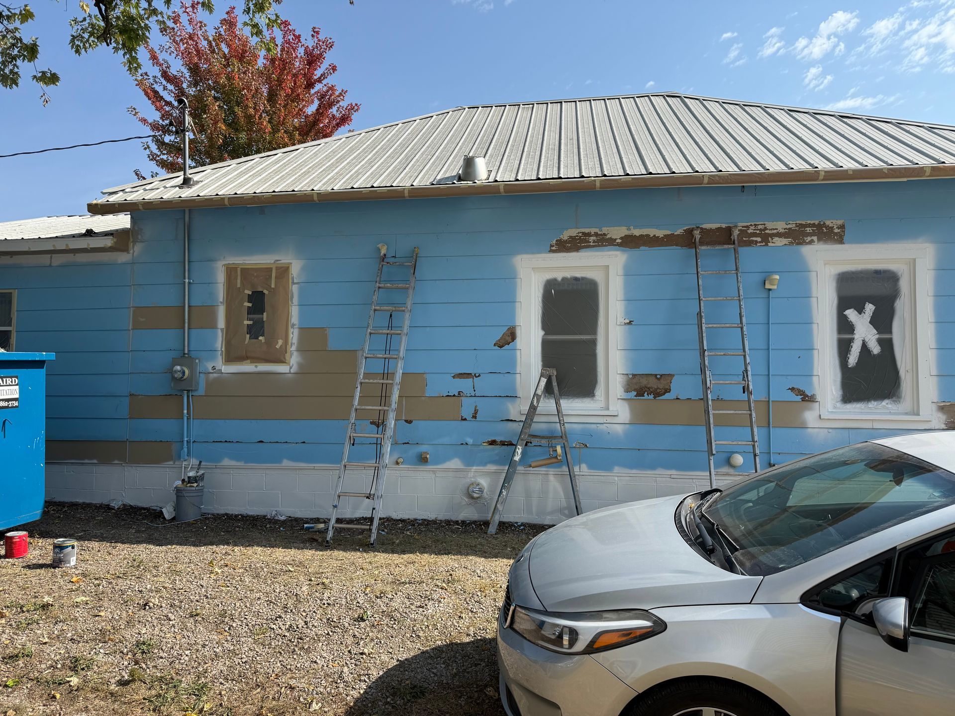 House exterior being painted, blue siding, ladders, silver car parked in front, dumpster on left.