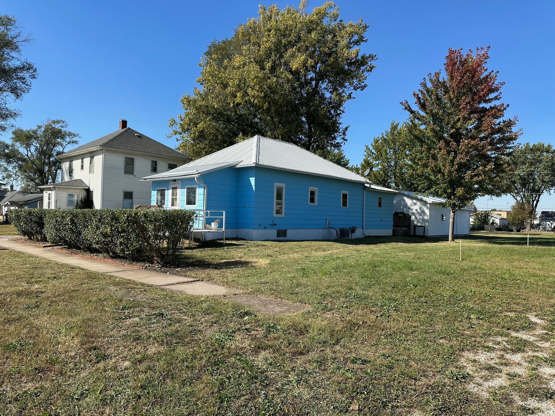 Blue house with white trim and metal roof, next to a white house, on a sunny, grassy lot.