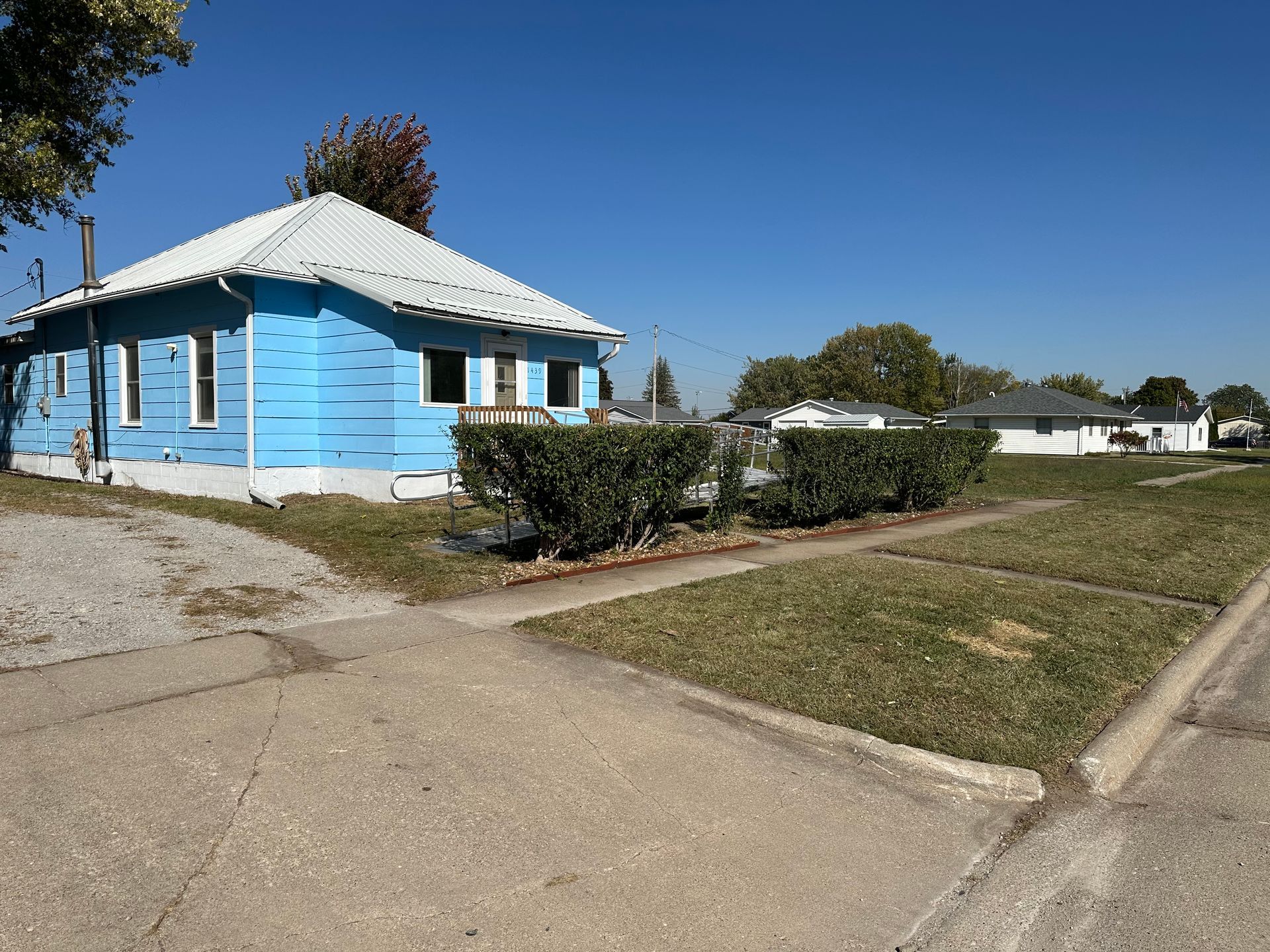Blue house with a corrugated metal roof, grass, bushes, and a paved street.