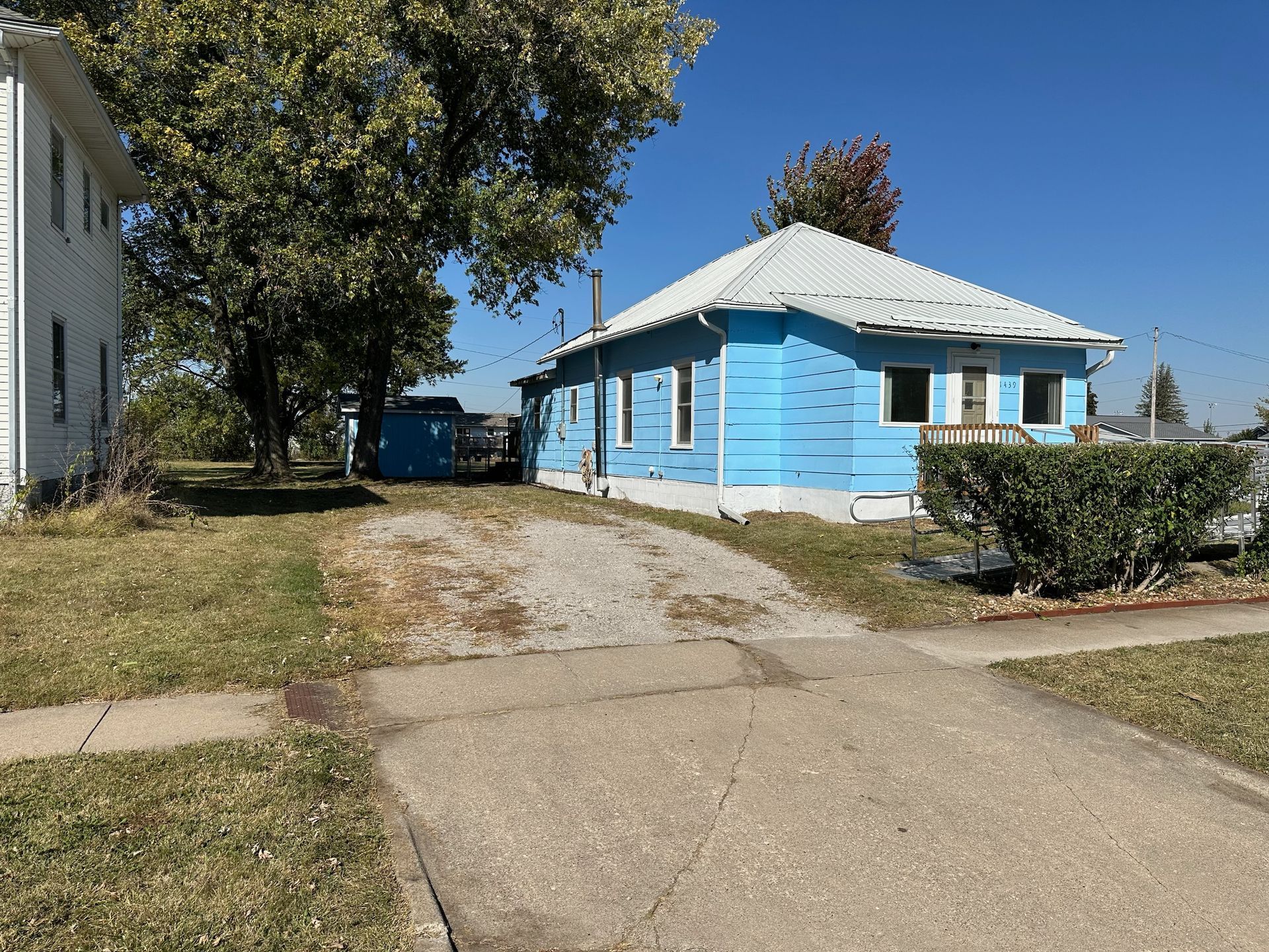 Blue-sided house with a gravel driveway and a large tree, next to a white house on a sunny day.