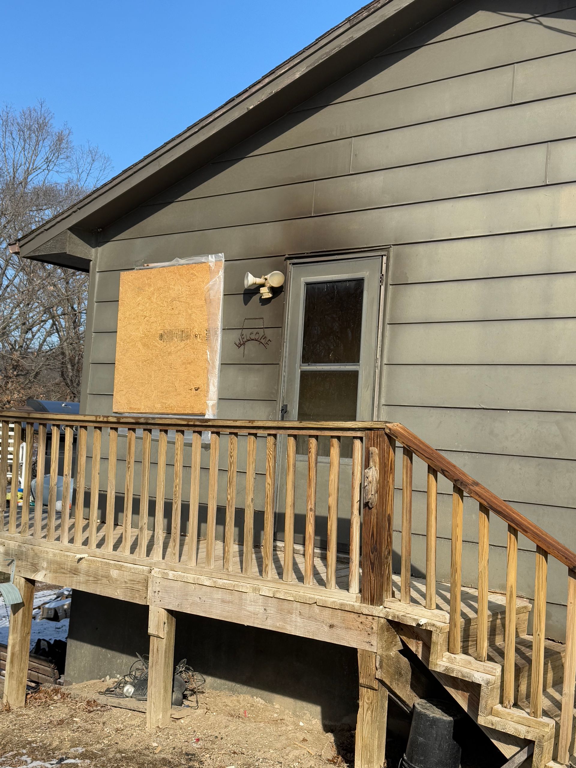 Exterior view of a house with smoke damage and a boarded-up window. A wooden deck and stairs lead to the door.