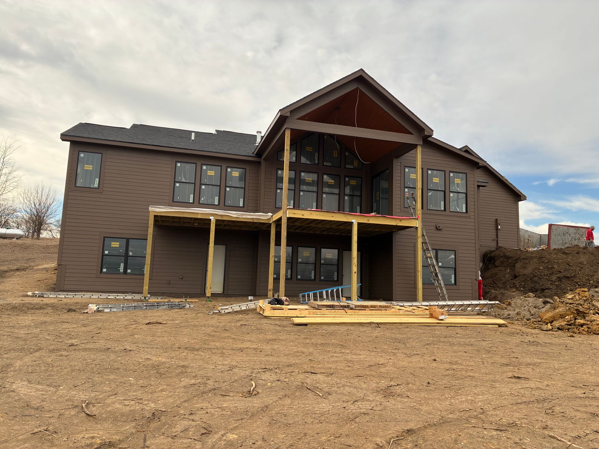 Brown two-story house under construction with a deck and large windows on a cloudy day.