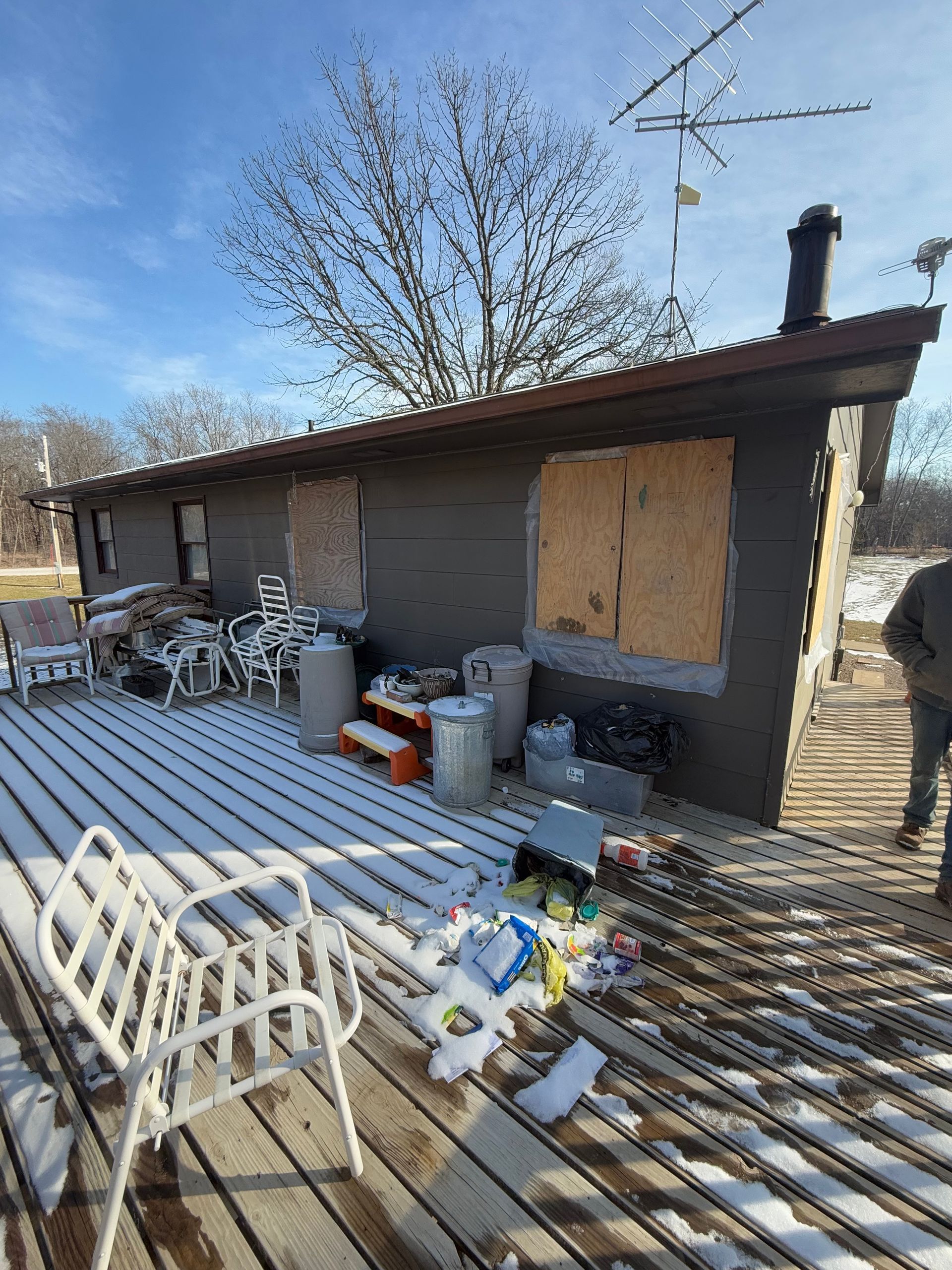 Exterior view of a weathered building with boarded-up window, cluttered deck covered in snow.