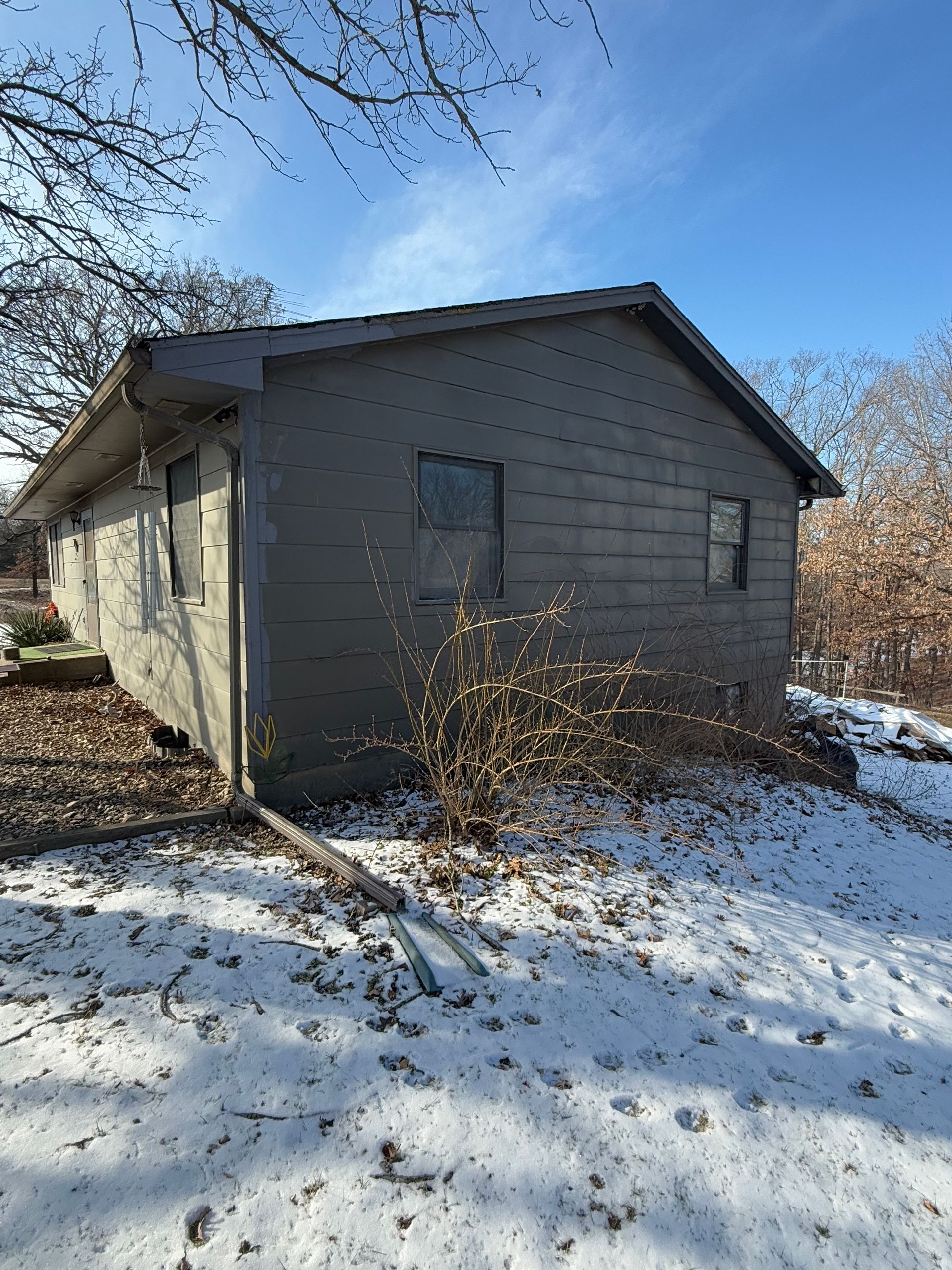 Gray house with two windows, covered in snow, surrounded by leafless trees under a blue sky.