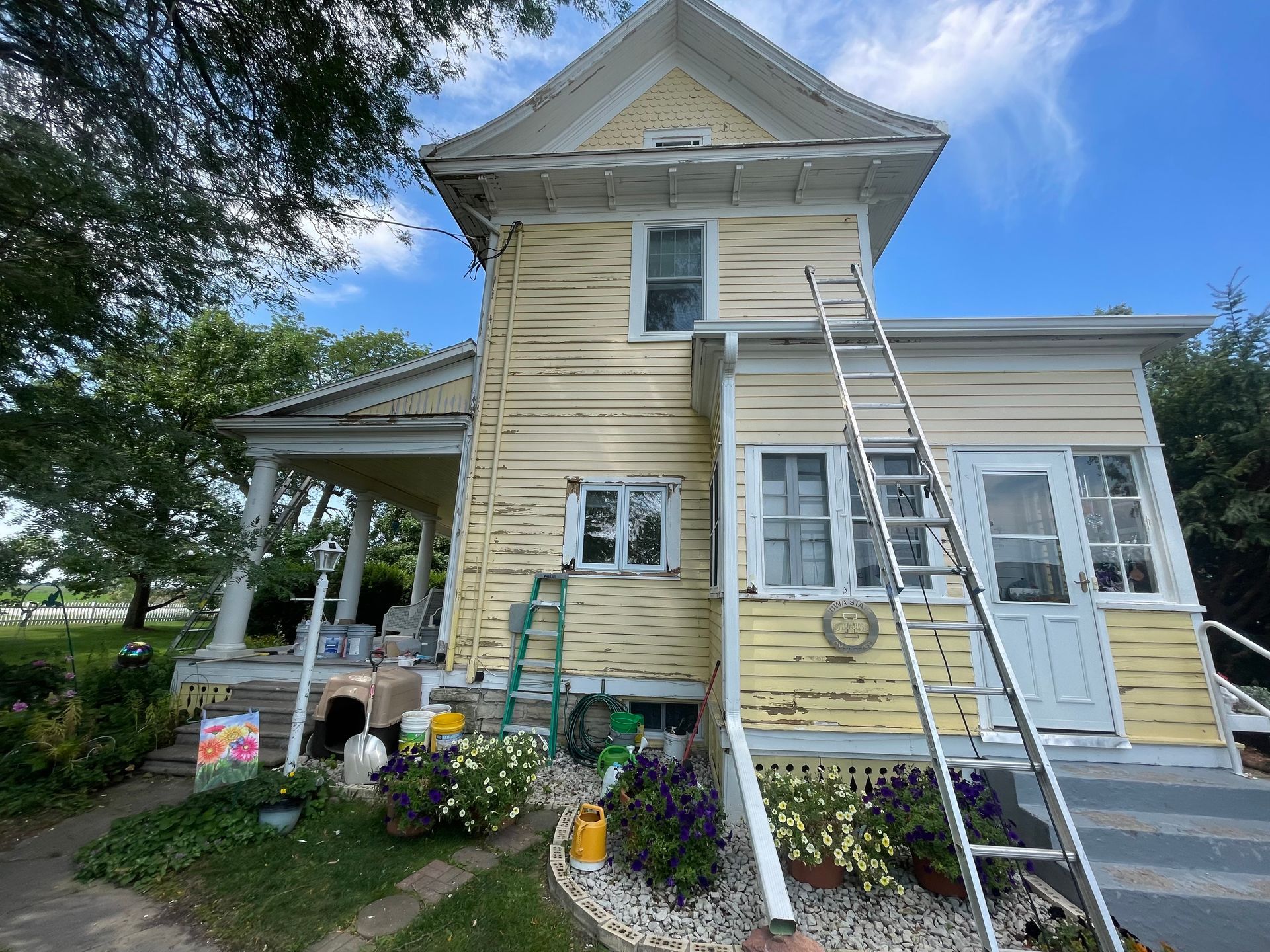 Two-story yellow house with porch. A ladder leans against the side, surrounded by painting supplies.