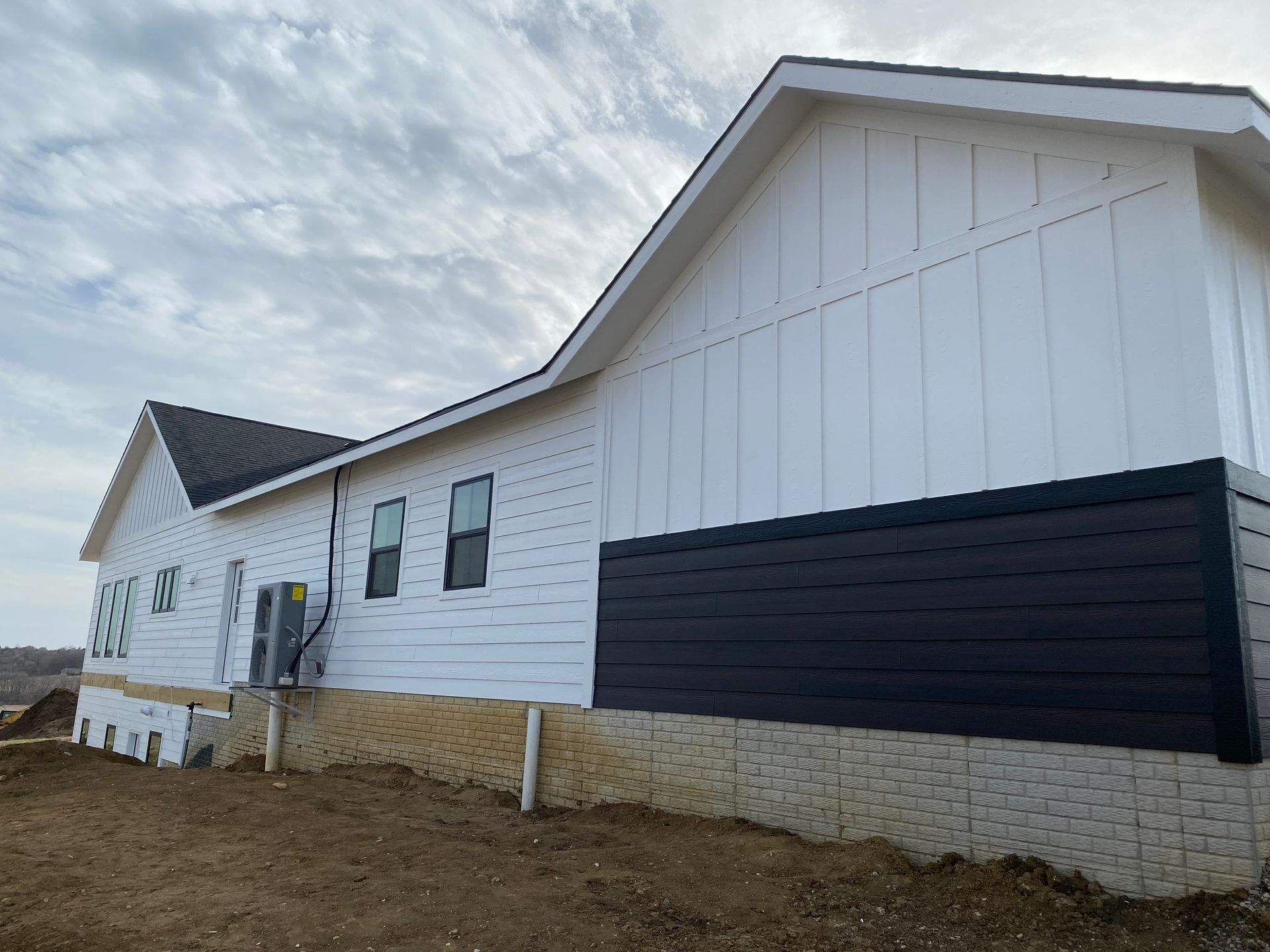 White house with black siding, windows, and partial exposed foundation. Cloudy sky background.